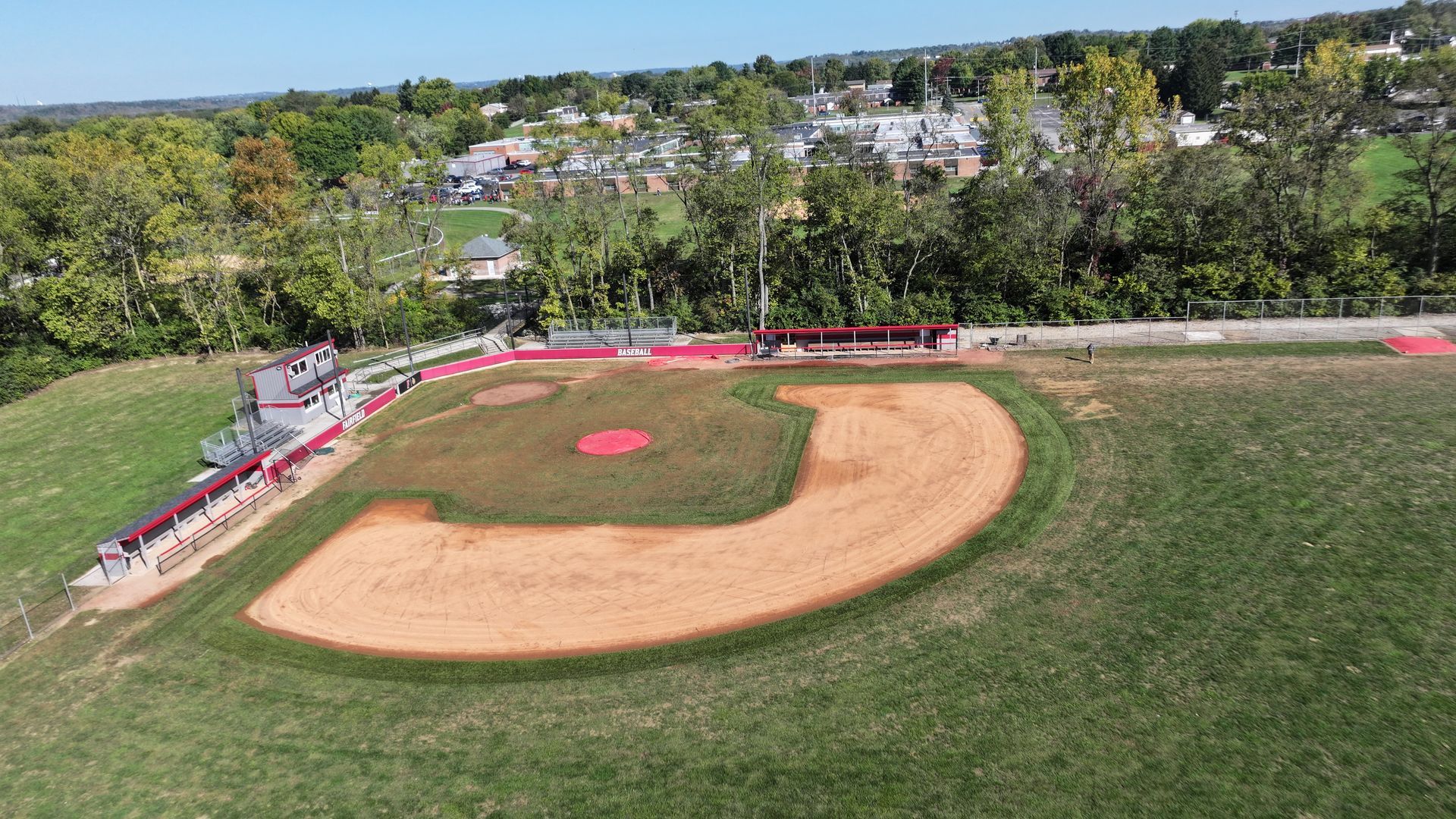 Aerial view of a baseball field with green grass, tan infield, and empty bleachers beside trees.