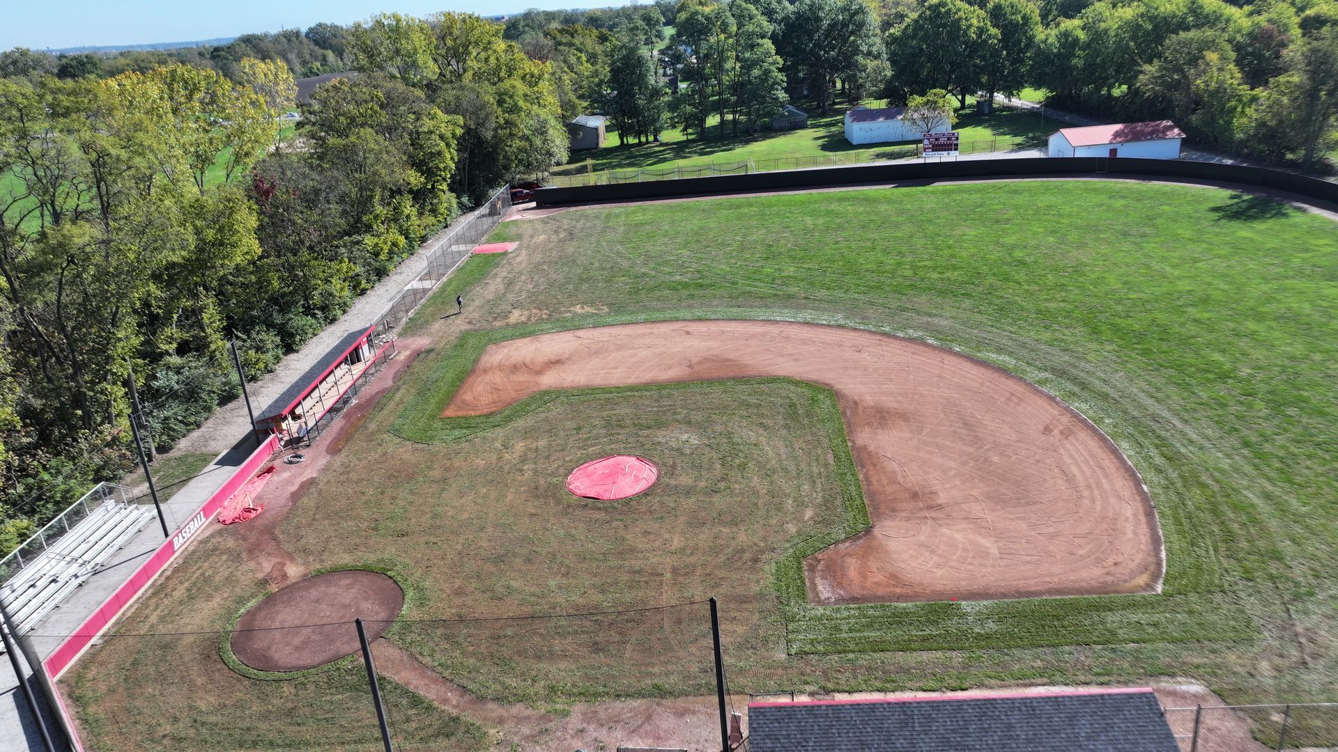 Aerial view of a baseball field with a dirt infield, green outfield, and trees beyond the fence