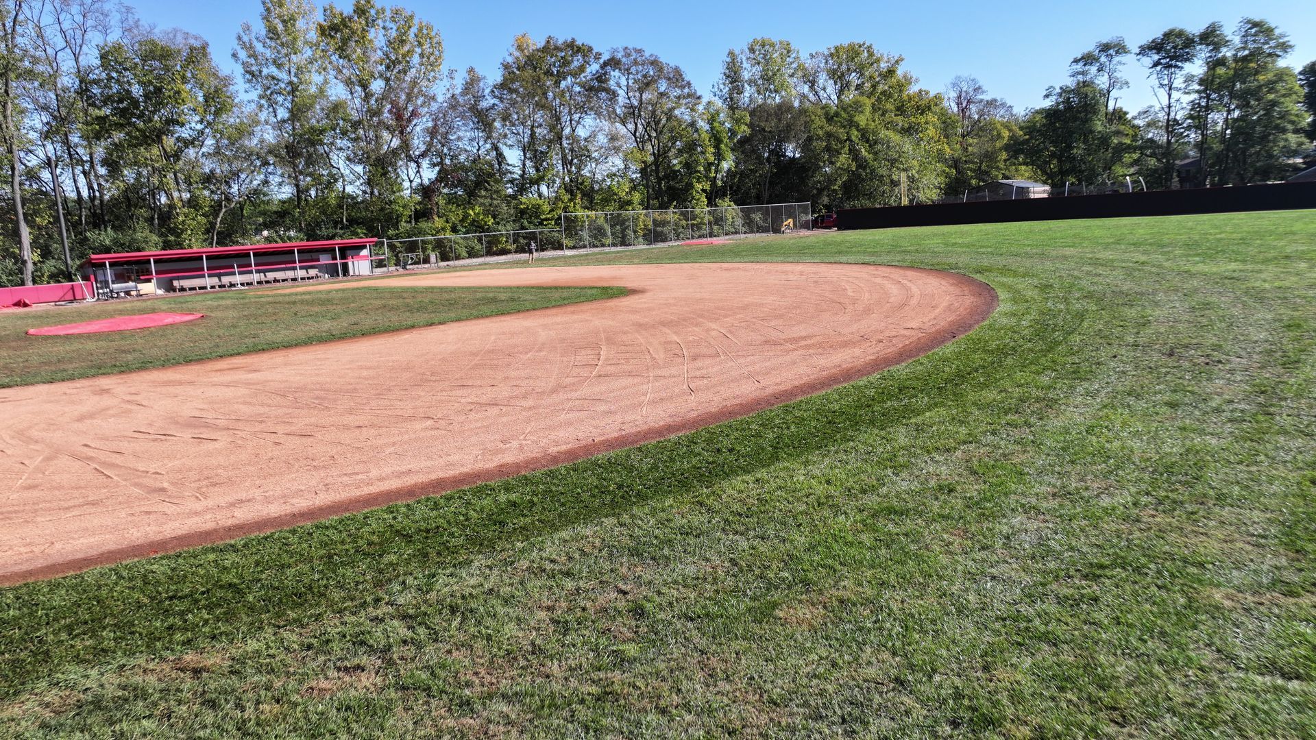 Baseball field with dirt infield, green outfield, and trees under a clear sky