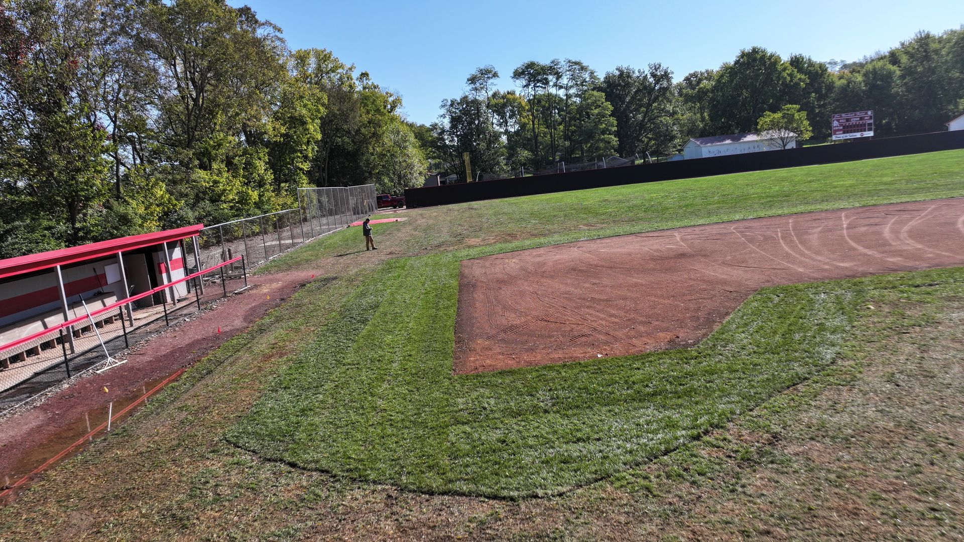 Baseball field with dirt infield, green grass, and red dugout fence under sunny skies