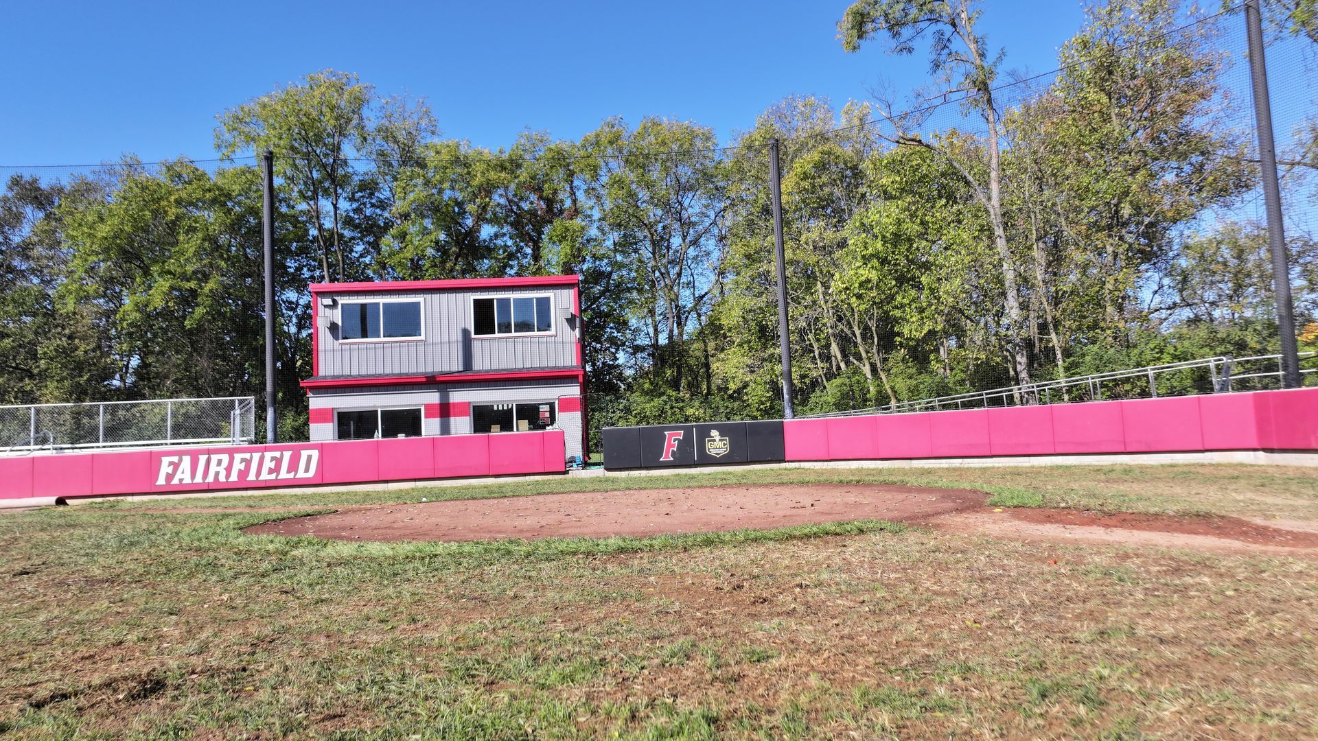 Baseball field with pink outfield wall, red press box, and trees under a clear blue sky