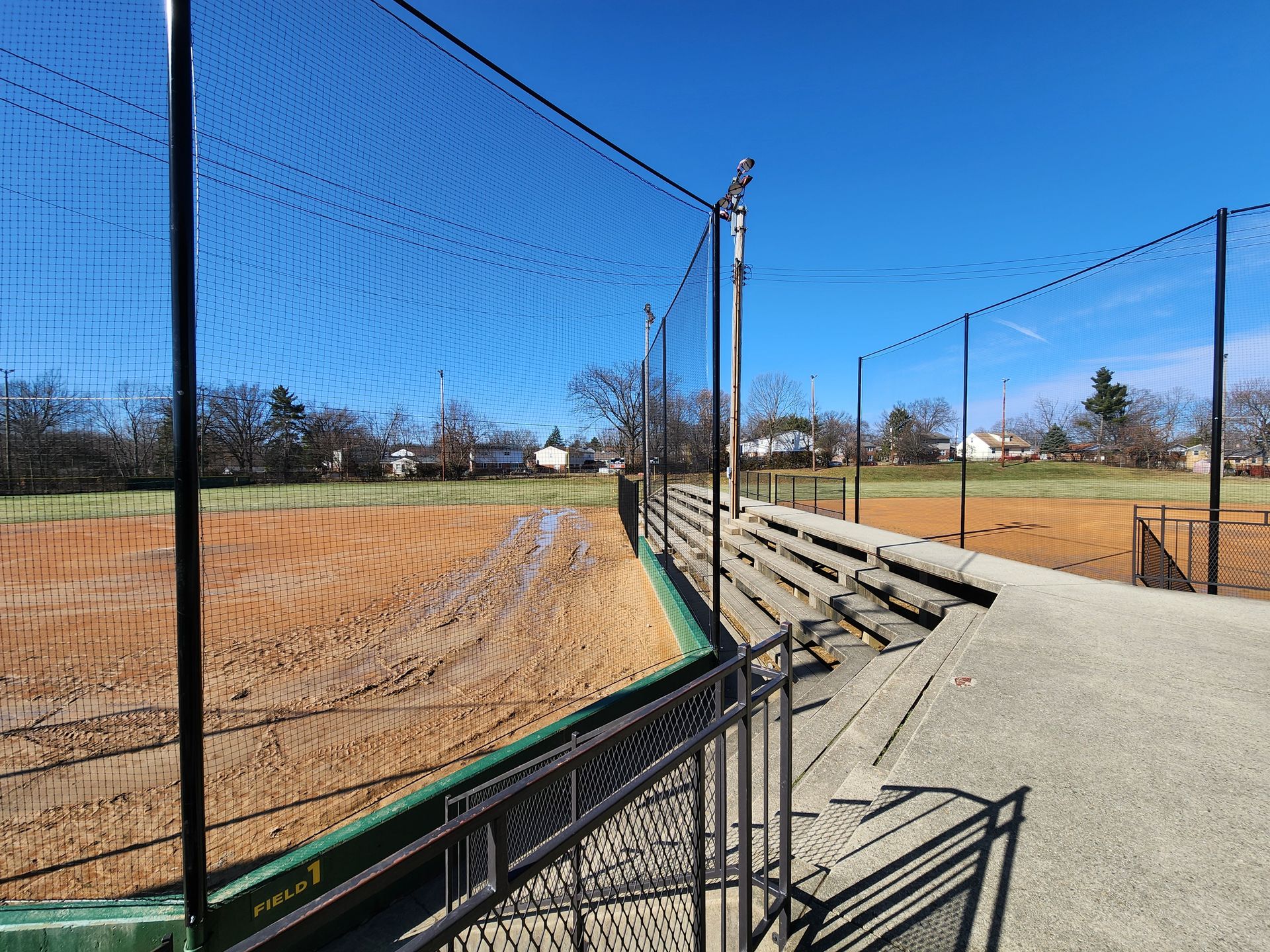 Empty baseball field with netting, bleachers, and a concrete walkway under a clear blue sky
