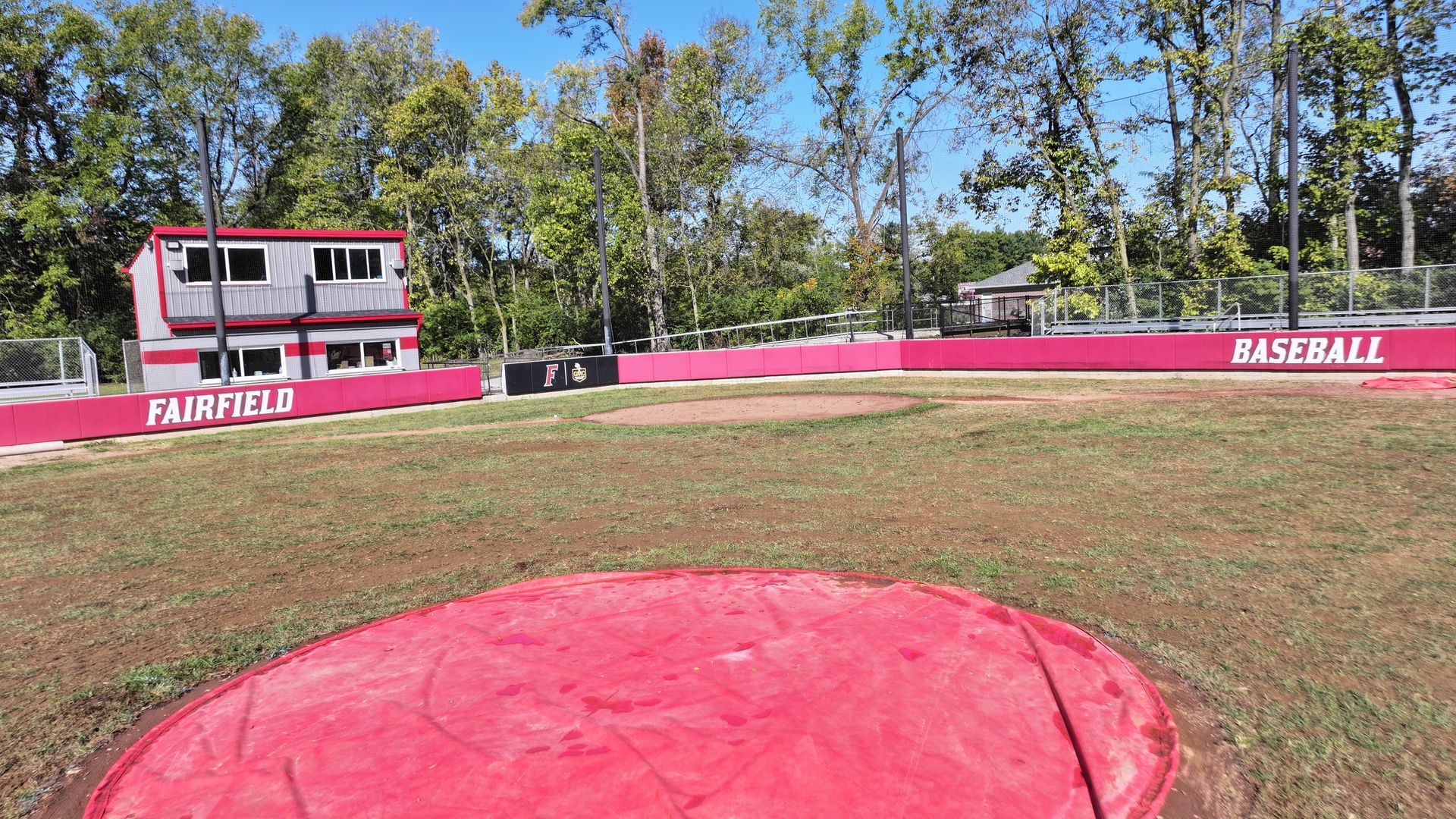 Empty baseball field with red infield, pink outfield fence, and scoreboard under a sunny sky