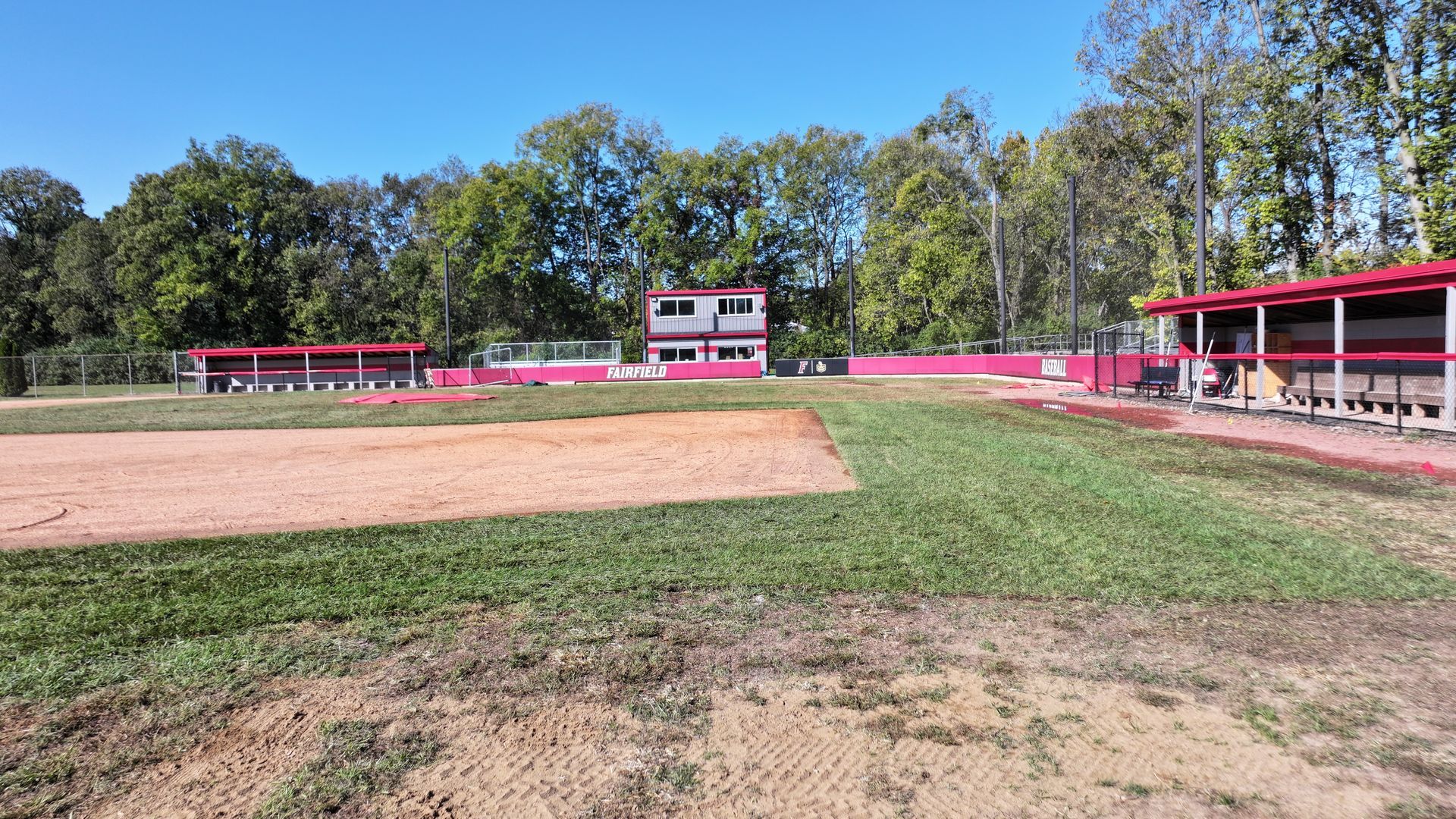 Baseball field with red bleachers, scoreboard, and trees under a clear blue sky