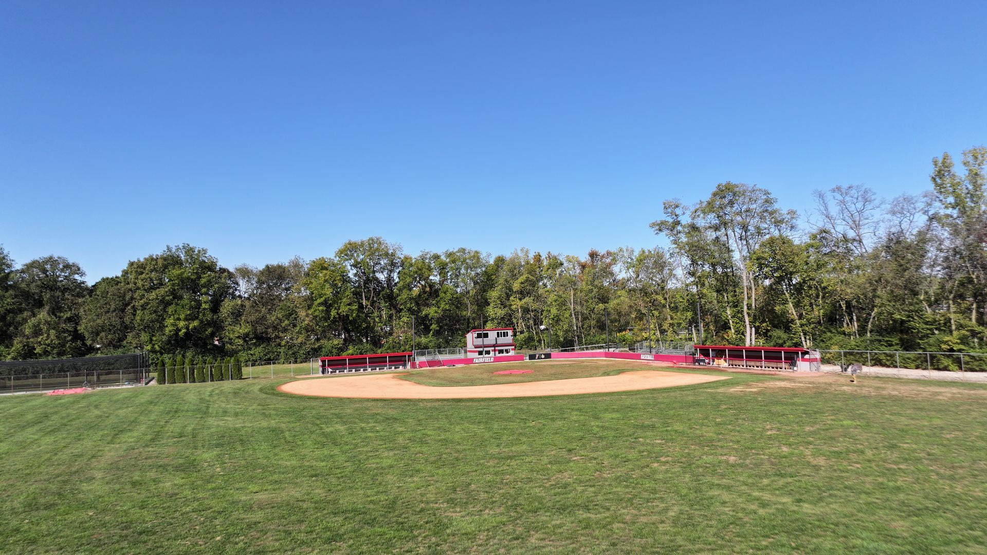 Baseball field with dirt infield, red benches, and trees under a clear blue sky.