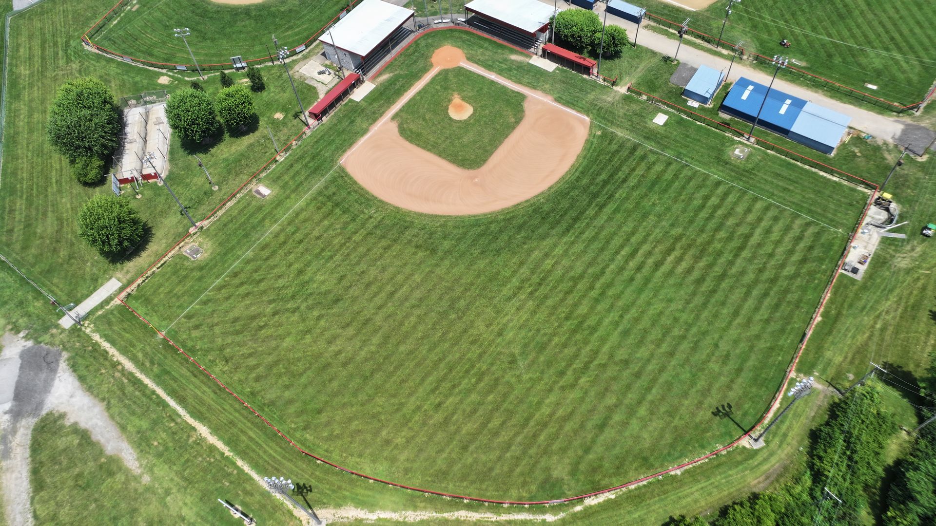 Aerial view of a baseball field with green grass, a dirt infield, and surrounding rural buildings.