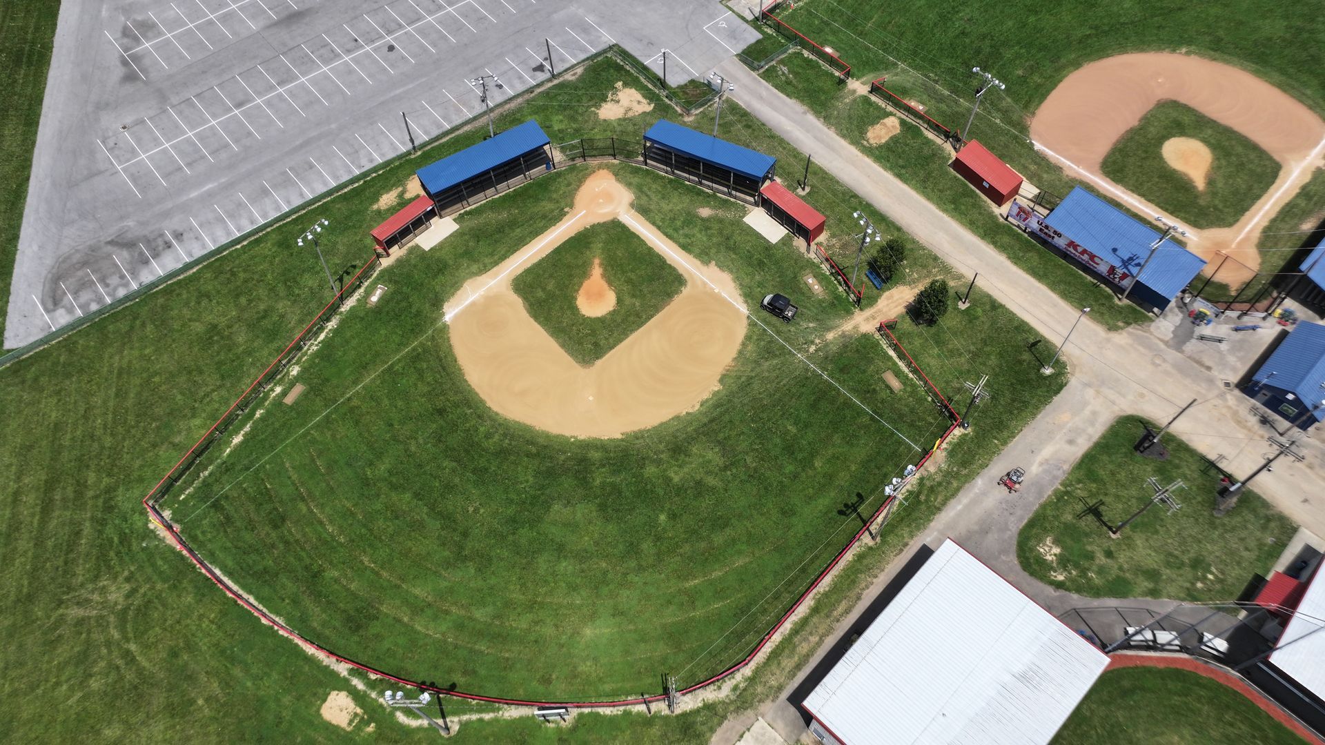 Aerial view of a baseball field with green outfield, dirt infield, dugouts, and nearby parking lots