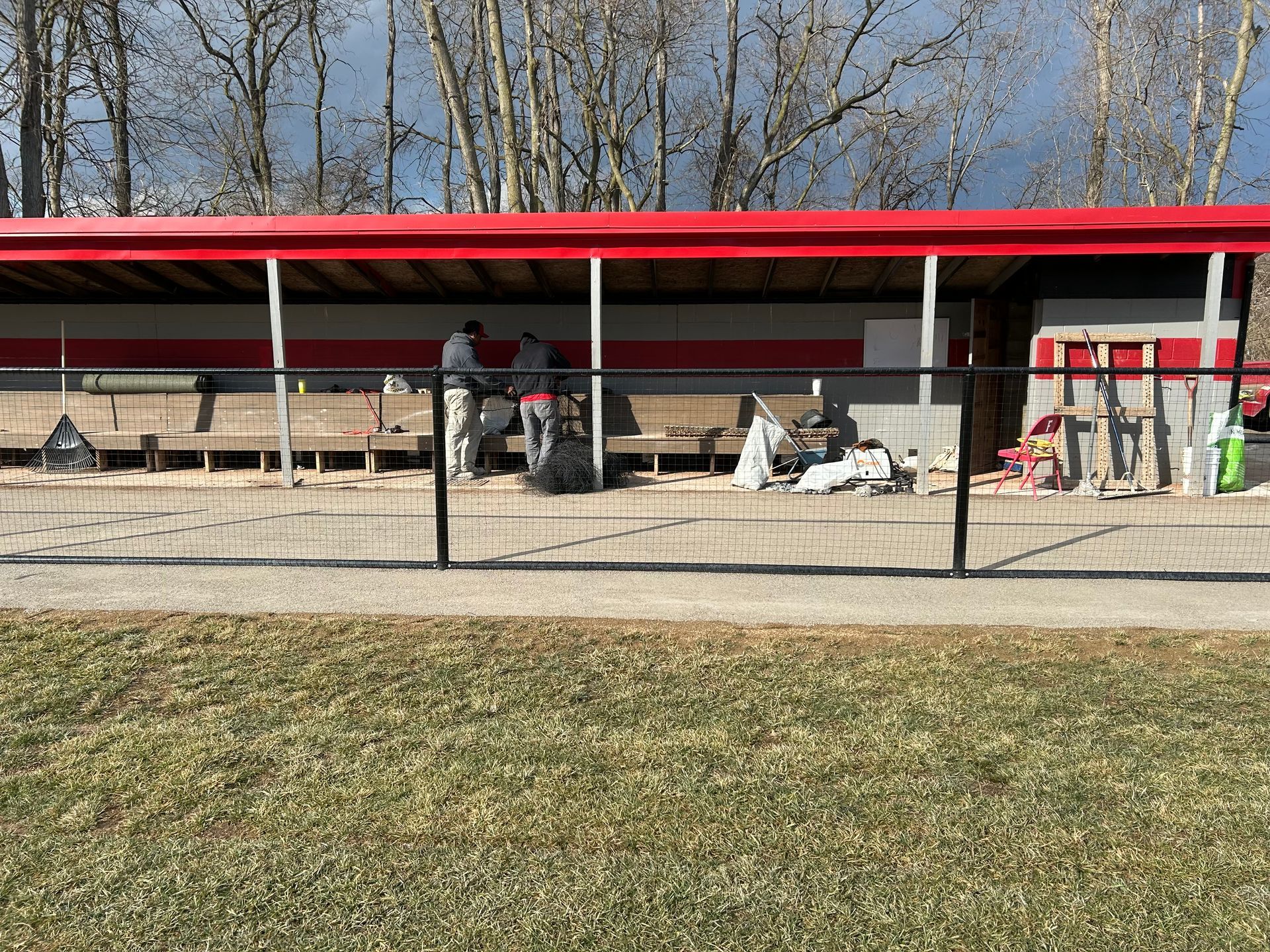 People standing under a red-roofed shelter by a fenced gravel area with equipment and trees behind.