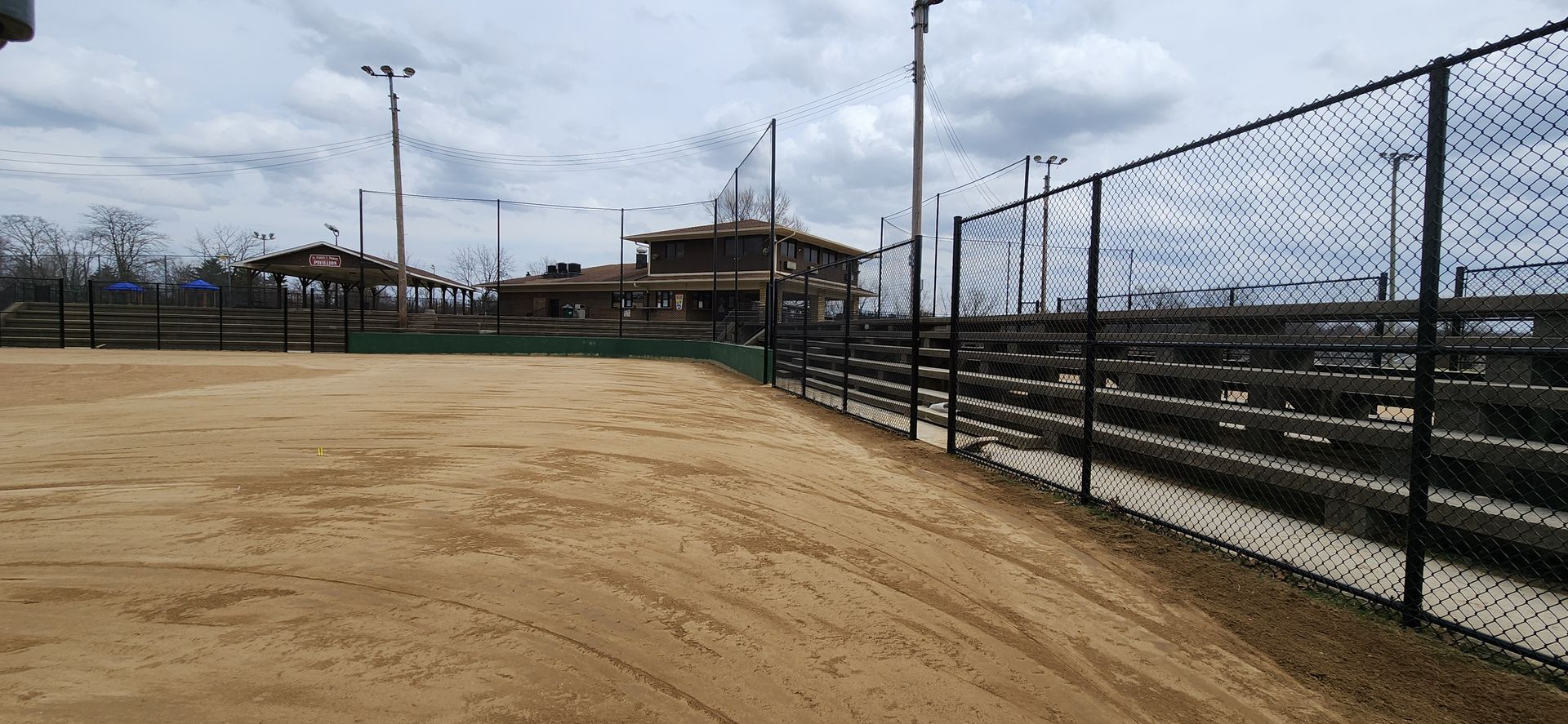 Empty dirt sports field beside a chain-link fence and bleachers under a cloudy sky