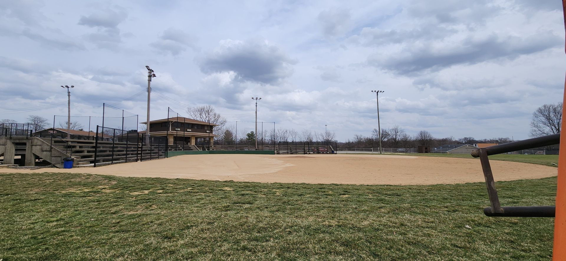 Empty baseball field under a cloudy sky with grass in the foreground and bleachers on the left