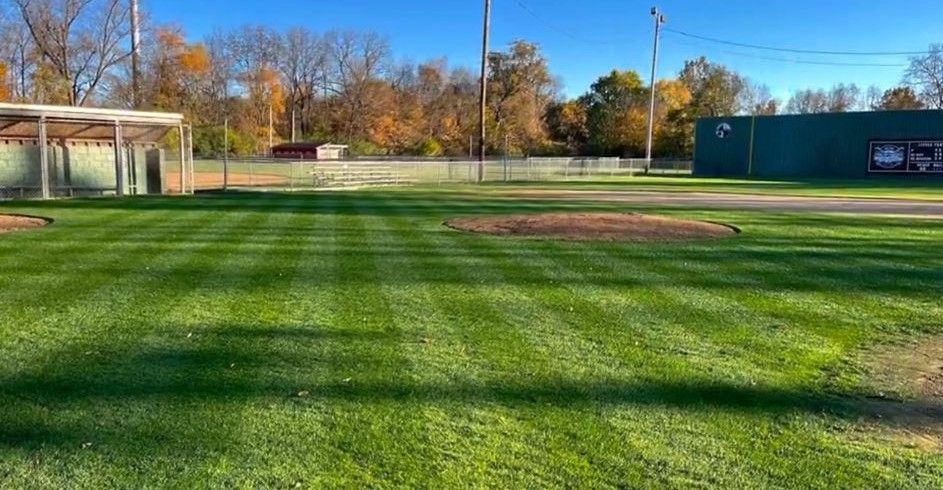 Empty grassy sports field with long shadows, trees, and a blue building in the background