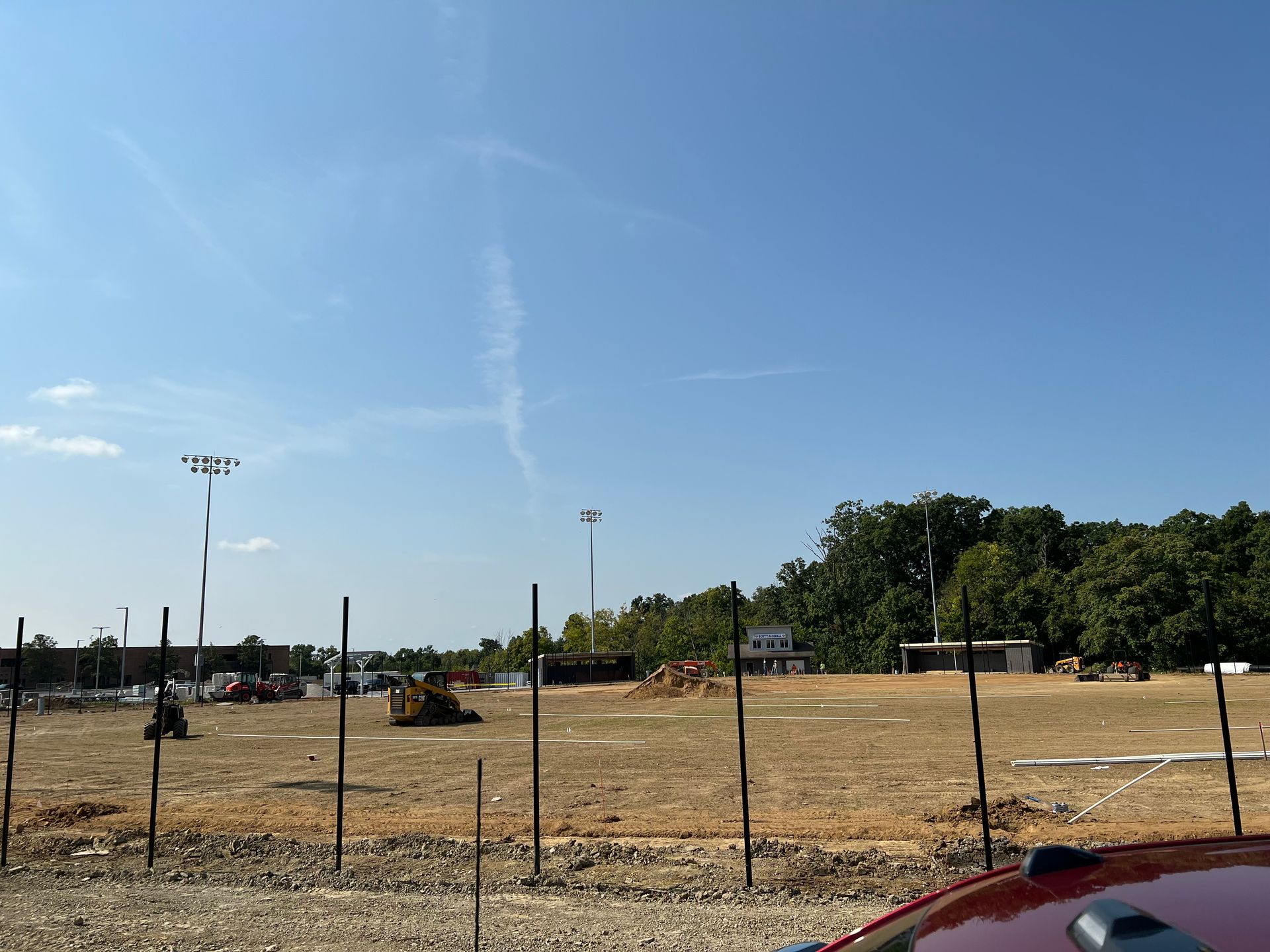 Open dirt field behind a fence under a clear blue sky, with a few light poles and trees in the distance