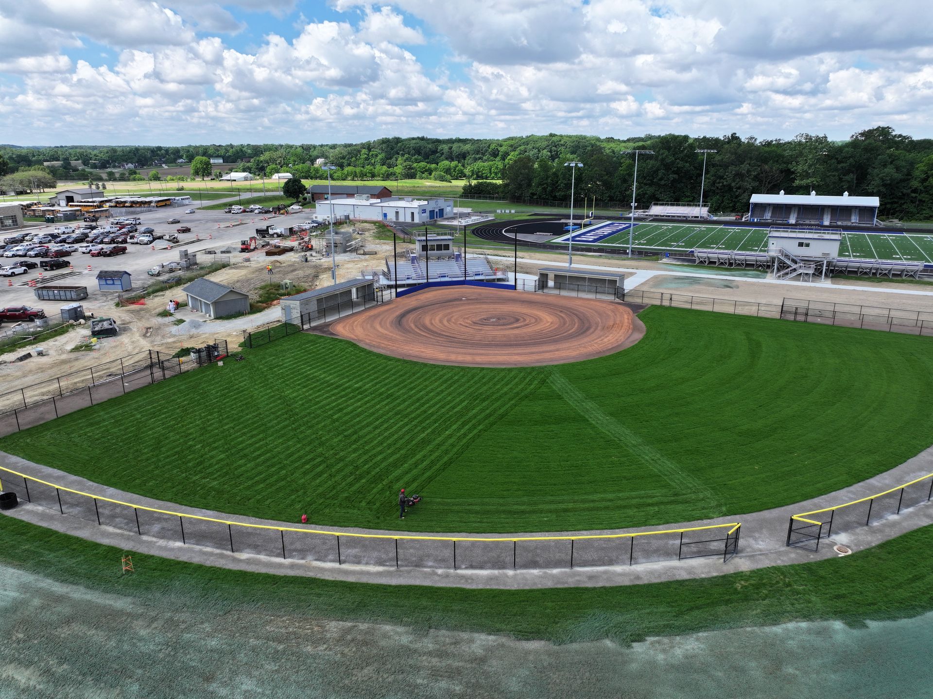 Aerial view of a green softball field with a red dirt infield under a cloudy sky.