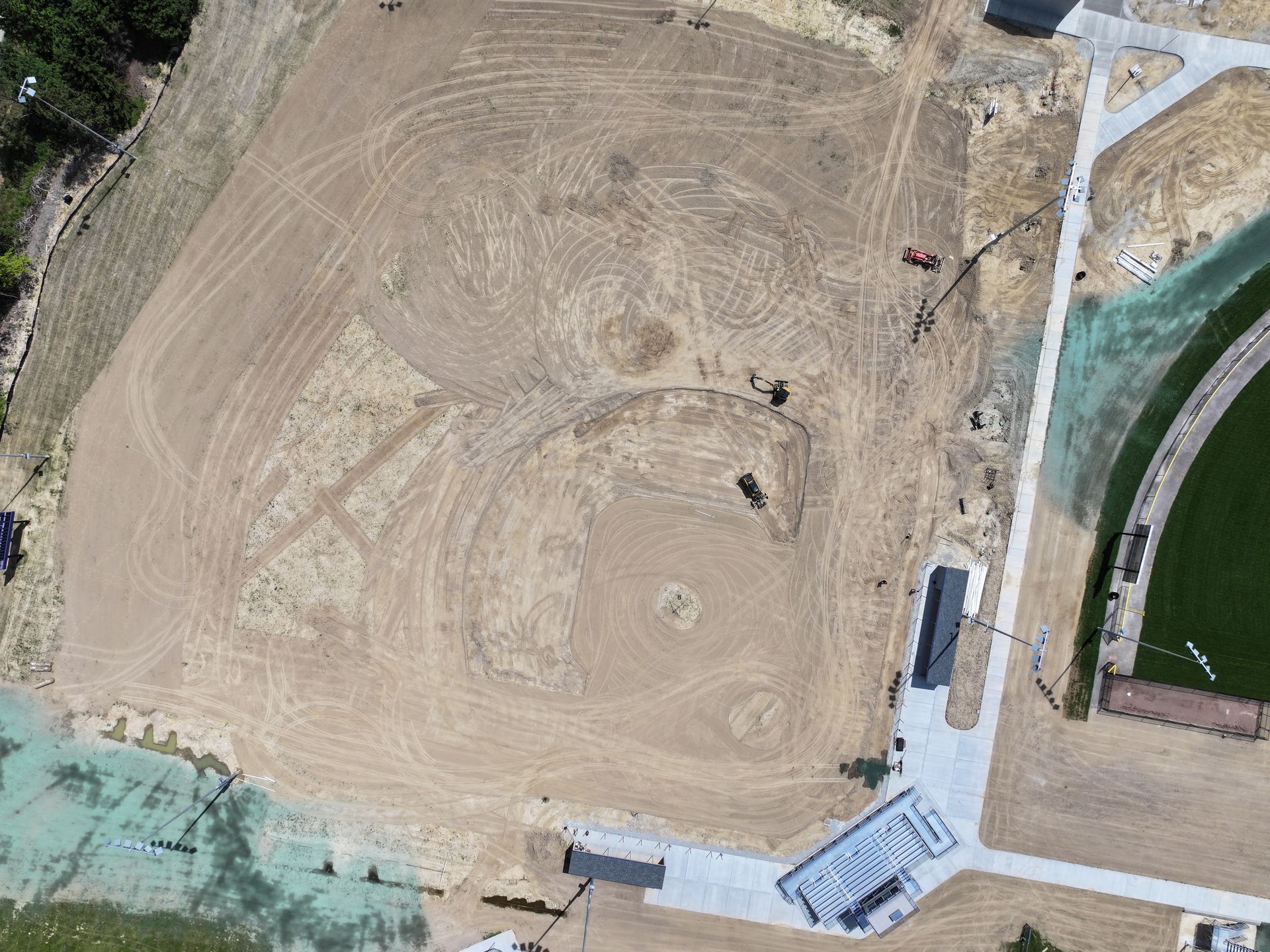 Aerial view of a large tan dirt construction site beside green sports fields and a paved area