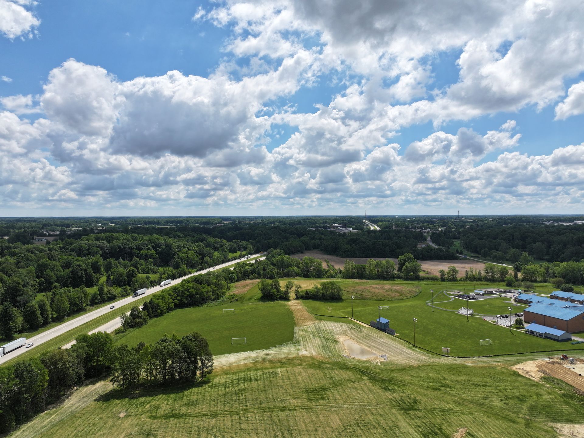 Wide rural landscape with green fields, a winding road, scattered buildings, and a cloudy blue sky