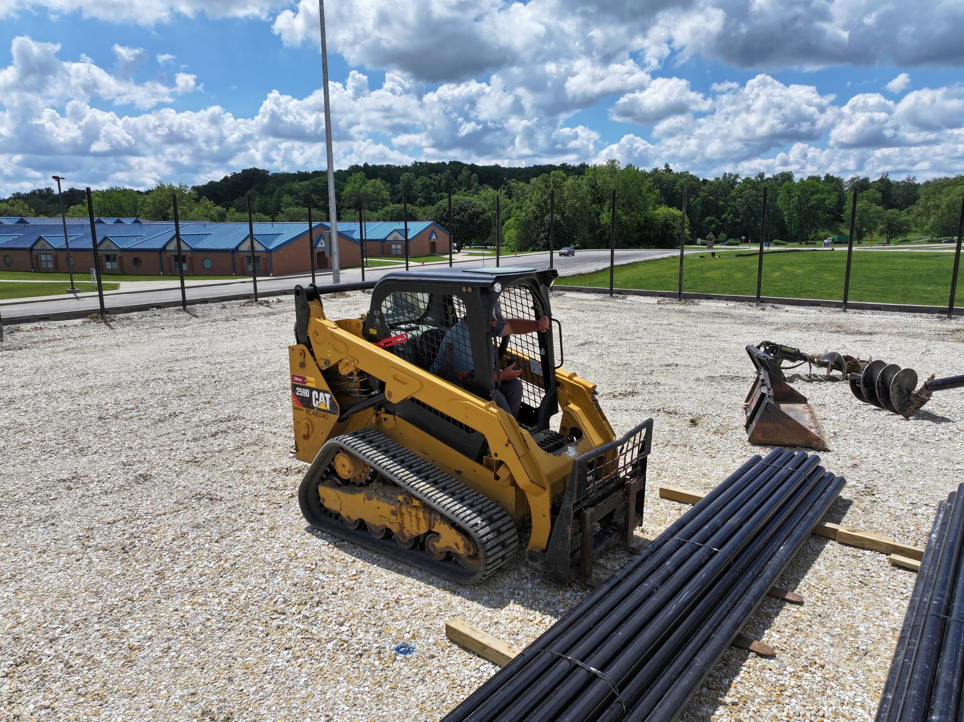 Yellow skid-steer loader on a gravel lot beside stacked black pipes under a cloudy sky