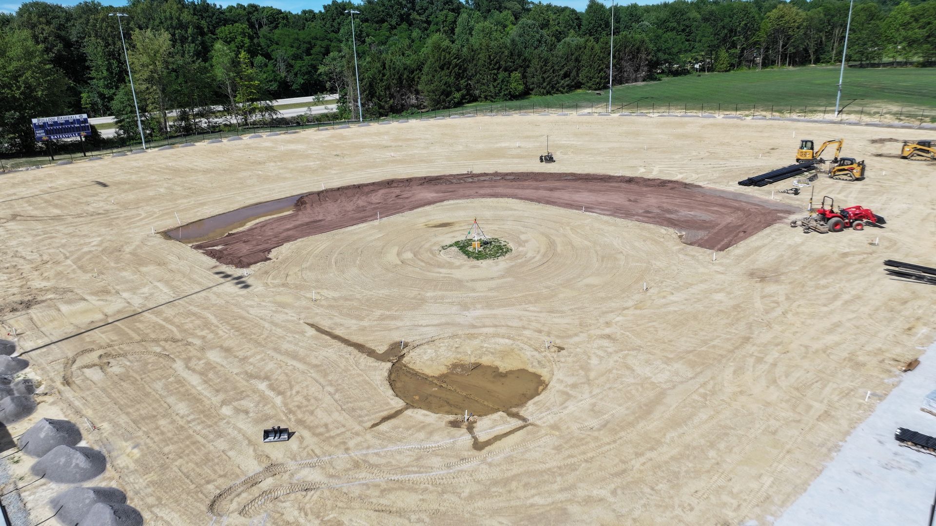 Construction site with a large circular dirt excavation, small machinery, and trees in the background
