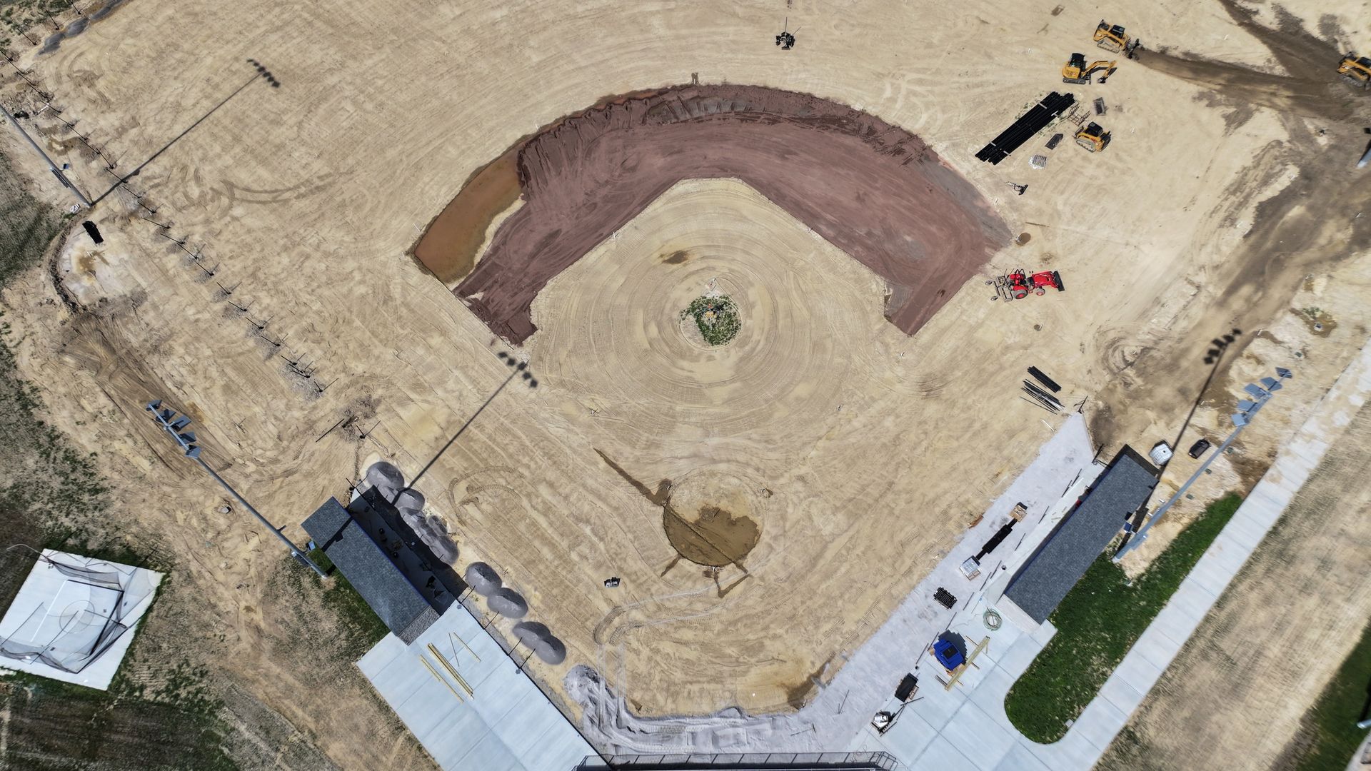 Aerial view of a baseball diamond under construction with dirt infield, dugouts, and surrounding pavement.
