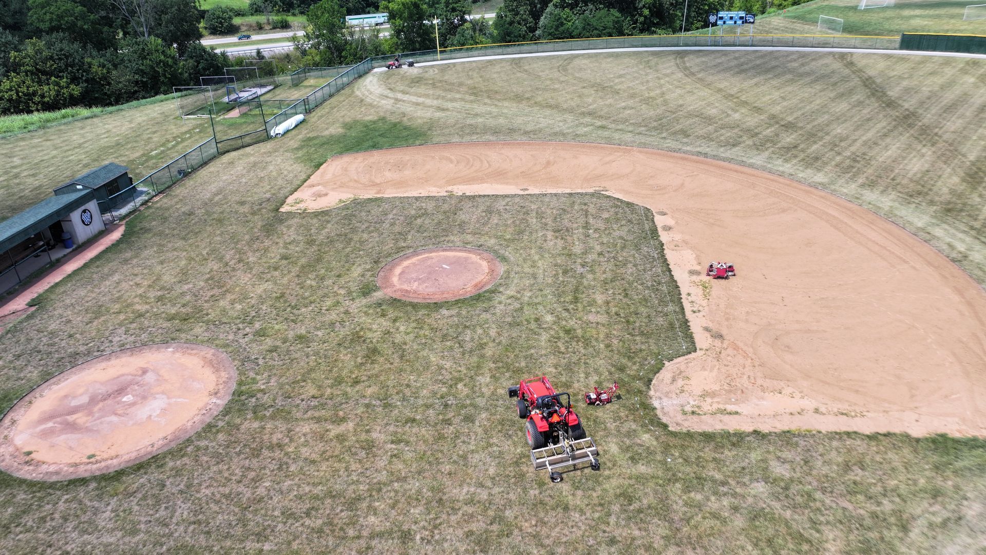 Aerial view of a baseball field with a dirt infield, grass outfield, and a red mower near home plate.