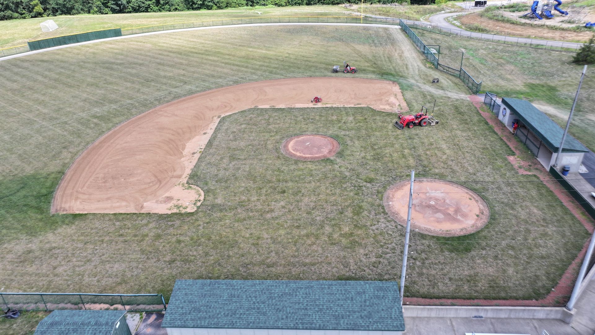Baseball field under construction with two red tractors and bare dirt infield areas