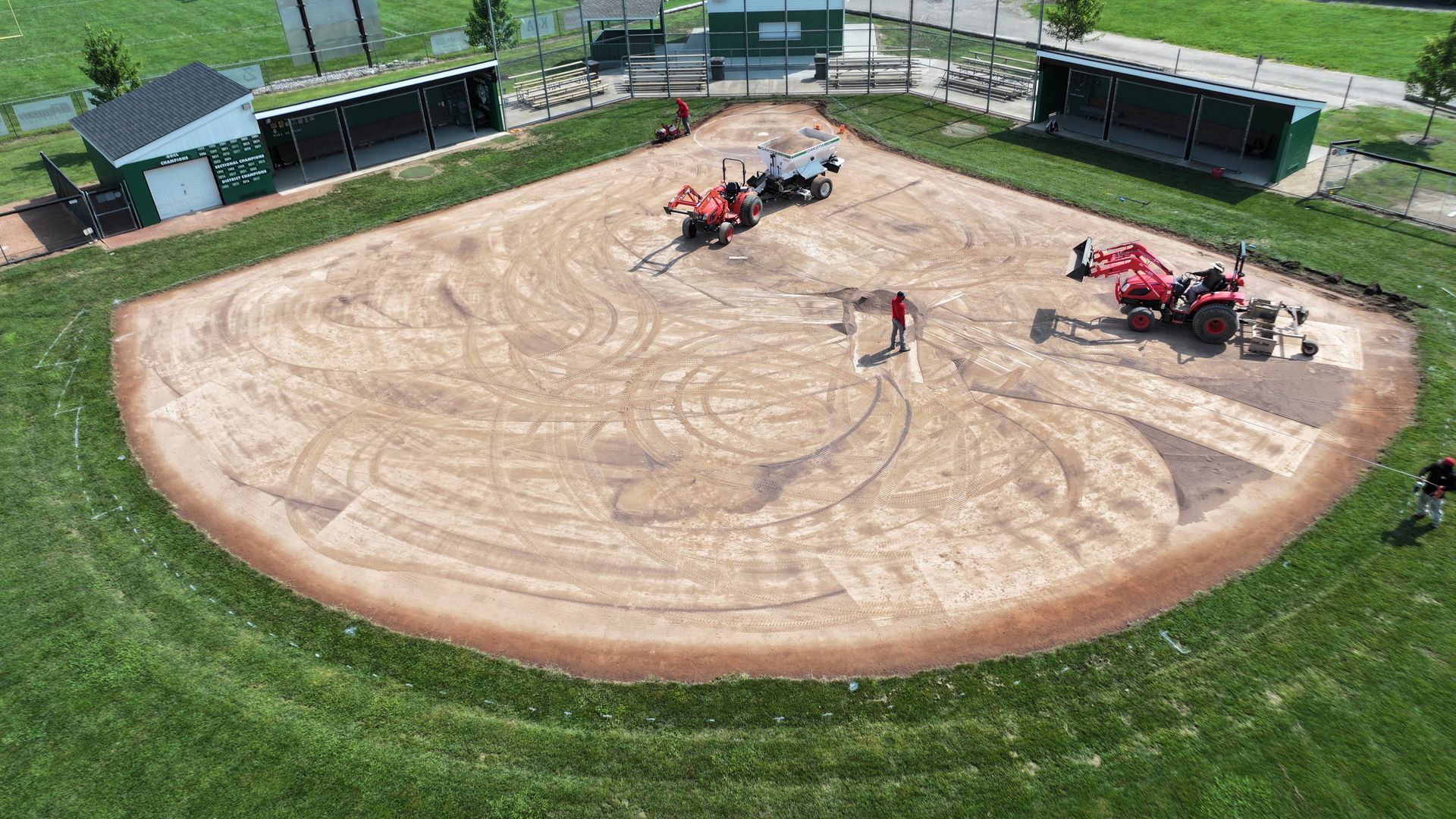 Aerial view of a sandy arena with two red tractors and workers inside a fenced grass field