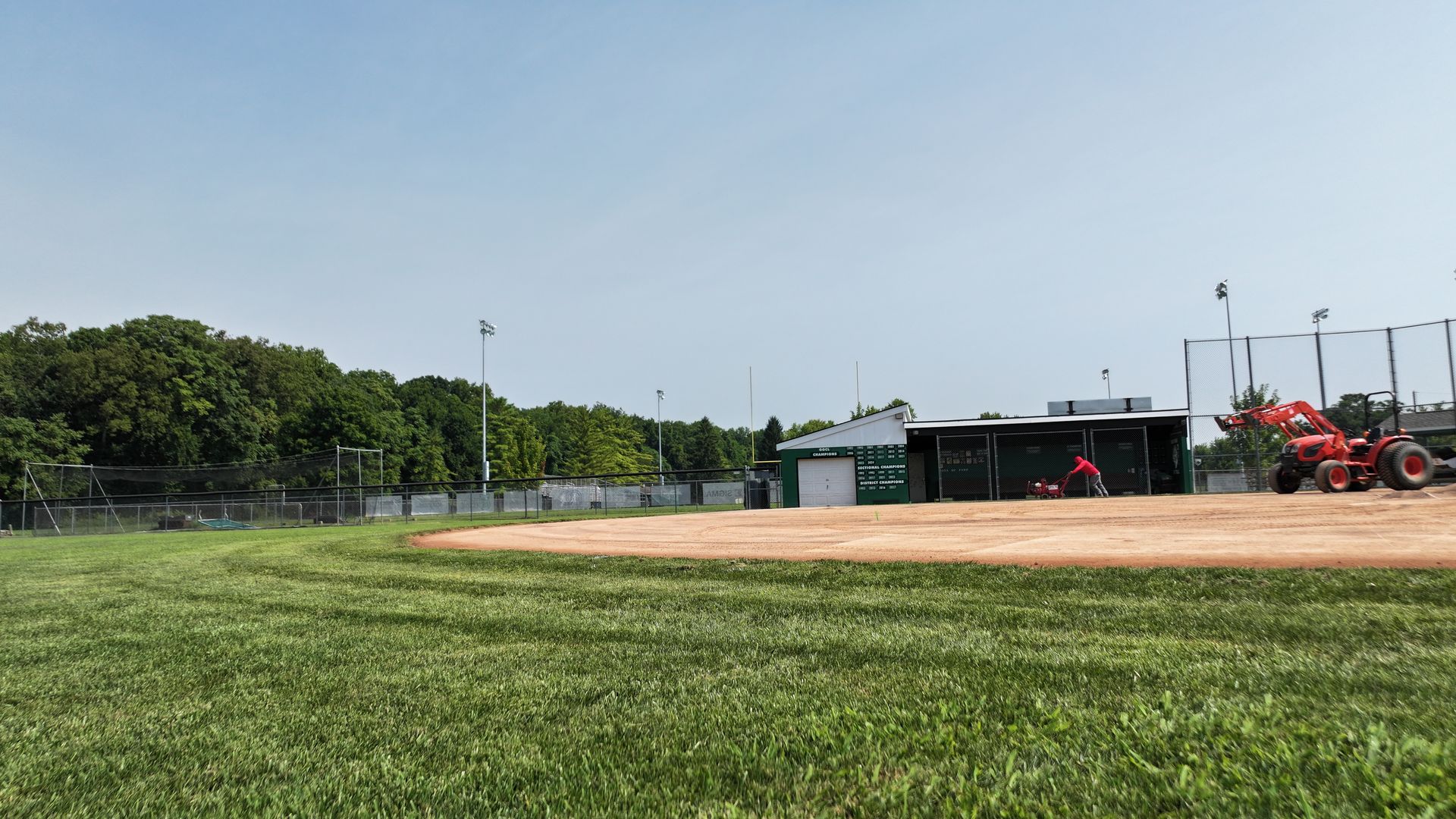 Baseball field with green grass, dirt infield, and players near the dugout on a sunny day