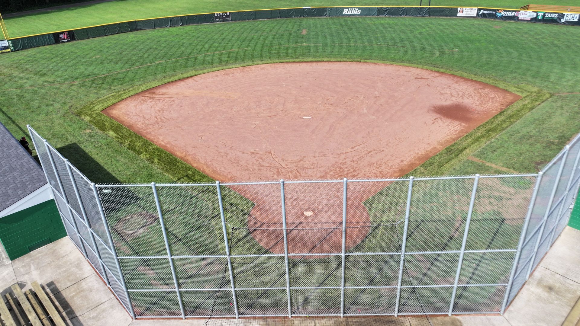 Empty baseball field viewed from behind a chain-link fence, with green outfield and brown infield dirt.