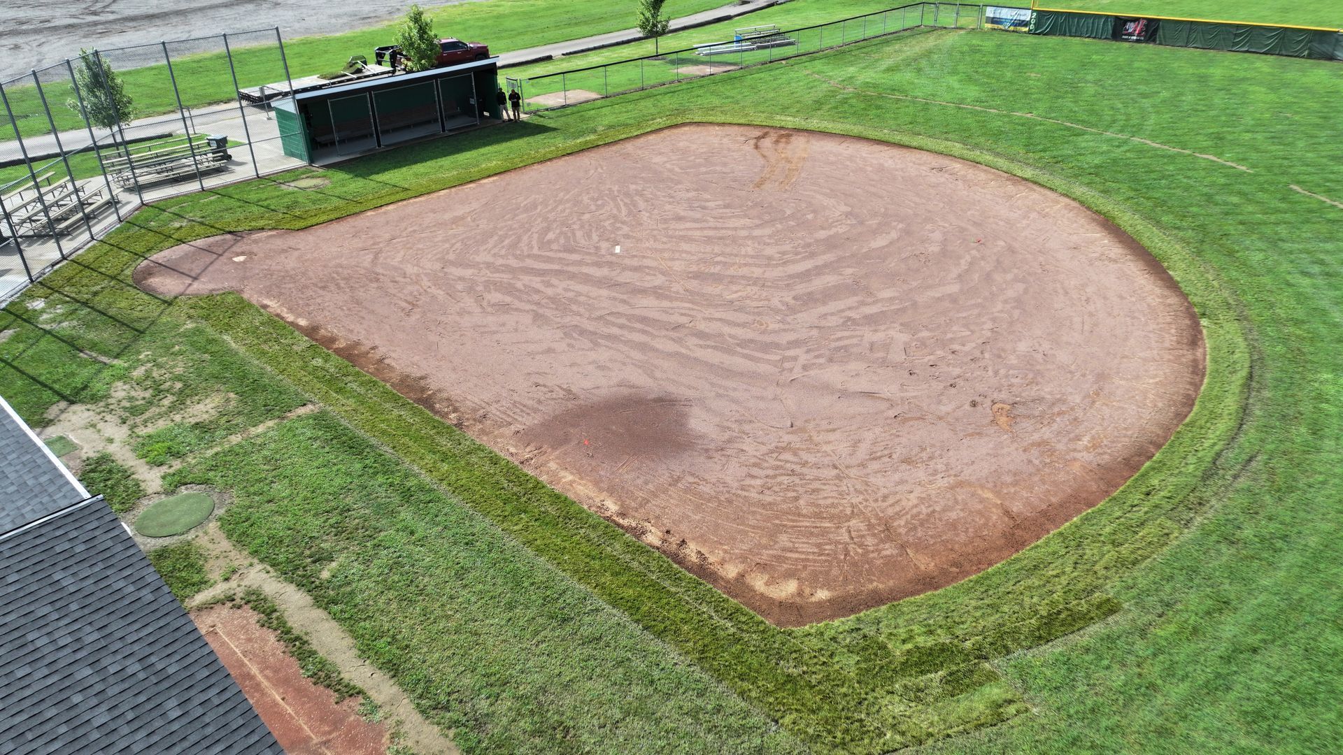 A dirt baseball field with green grass surrounding it and a small dugout in the background