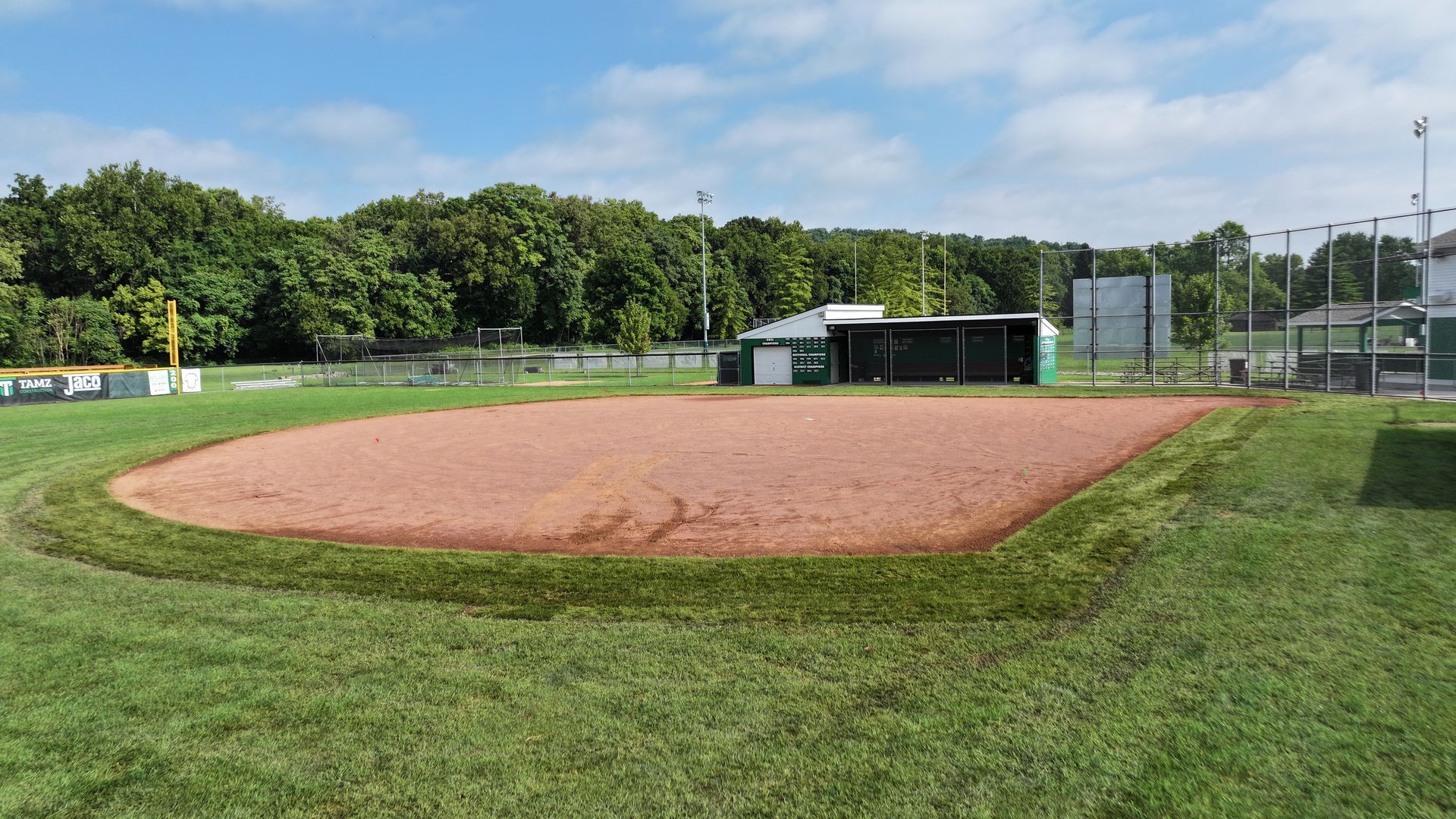 Empty baseball field with dirt infield, green grass, dugout, and trees in the background