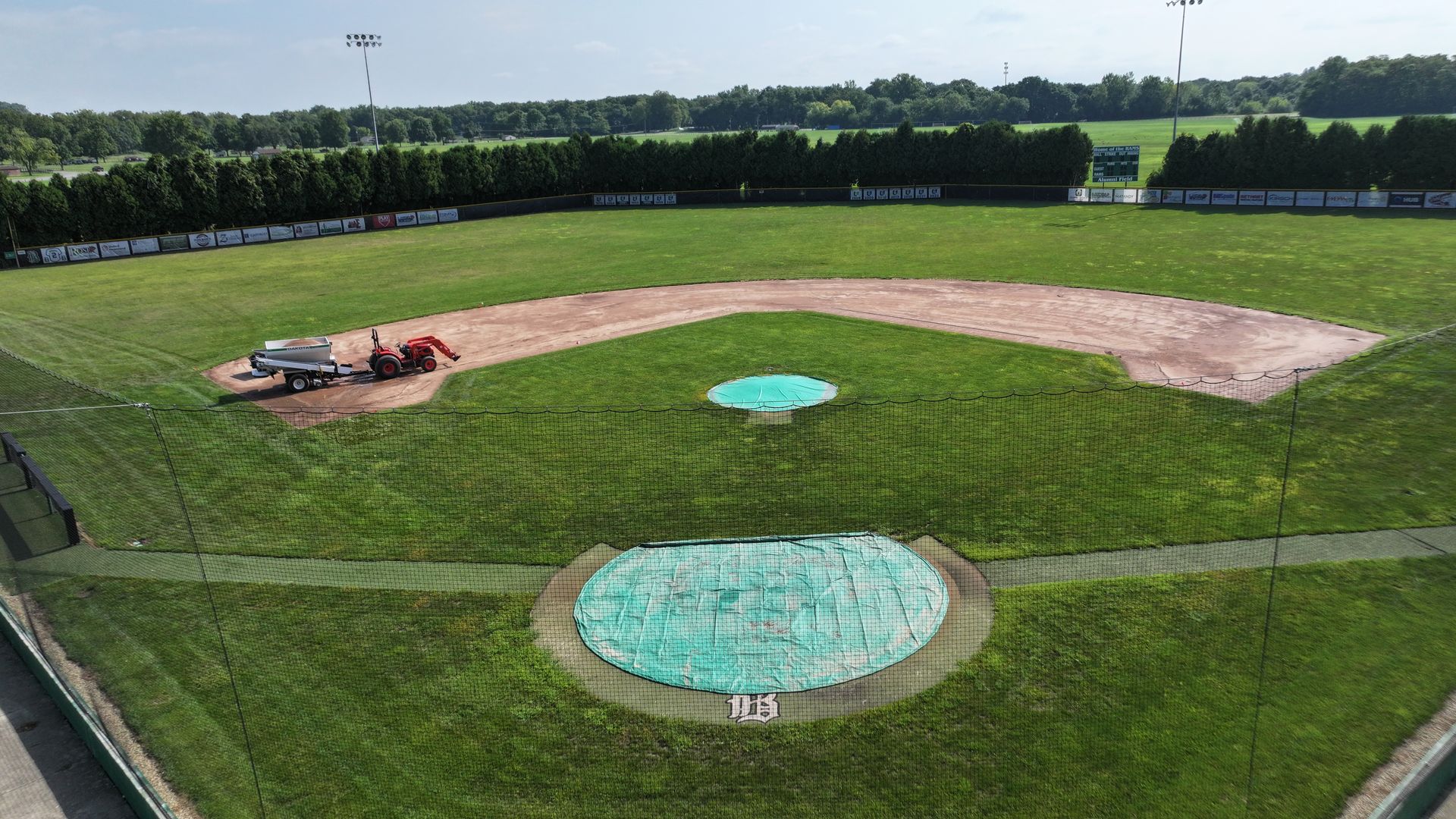 Baseball field with a red groundskeeper cart on the left and blue tarps on the infield, seen from above