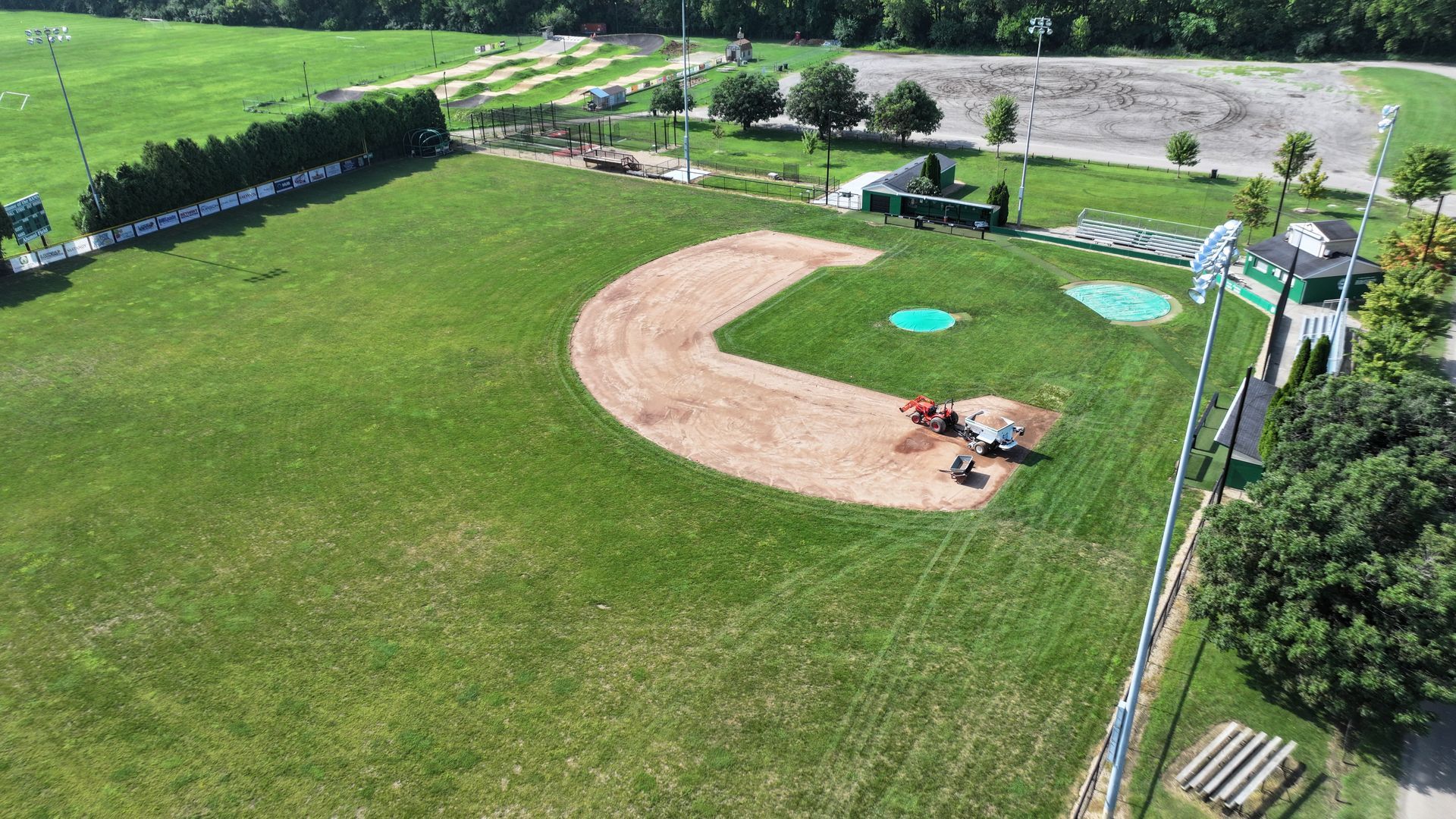 Aerial view of a baseball field beside a river, with green grass, infield dirt, and a few people near the dugout.