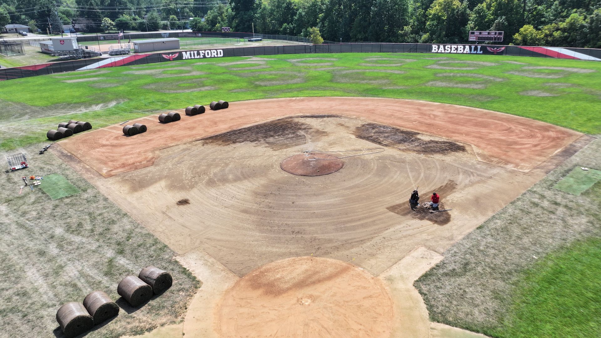 Aerial view of a baseball field with a dirt infield, green outfield, and a few people near home plate.