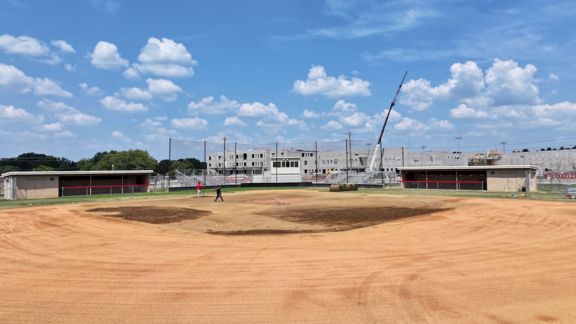 Wide dirt field with low buildings and a crane under a bright blue sky with scattered clouds