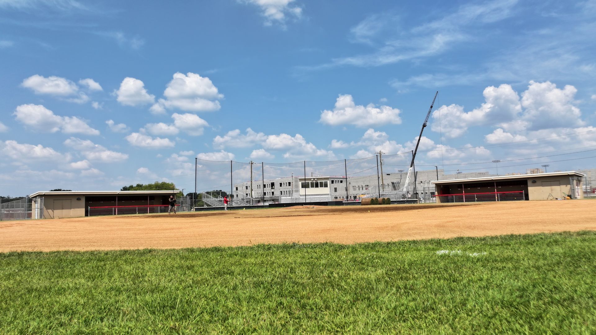 Wide grassy field in foreground with a low building and crane under a blue, partly cloudy sky