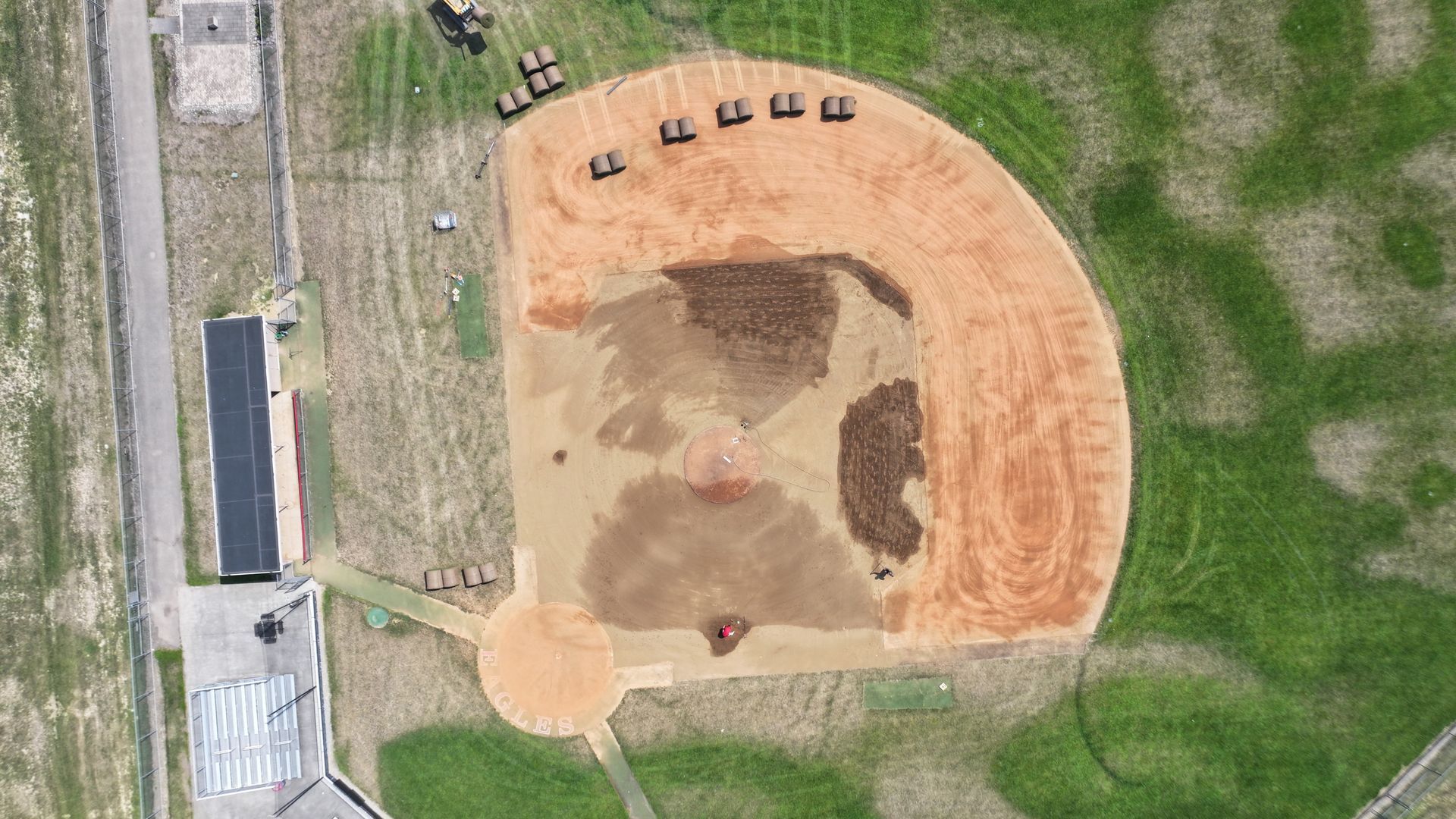Aerial view of a dirt sports field with a small dugout and benches beside a grassy area.