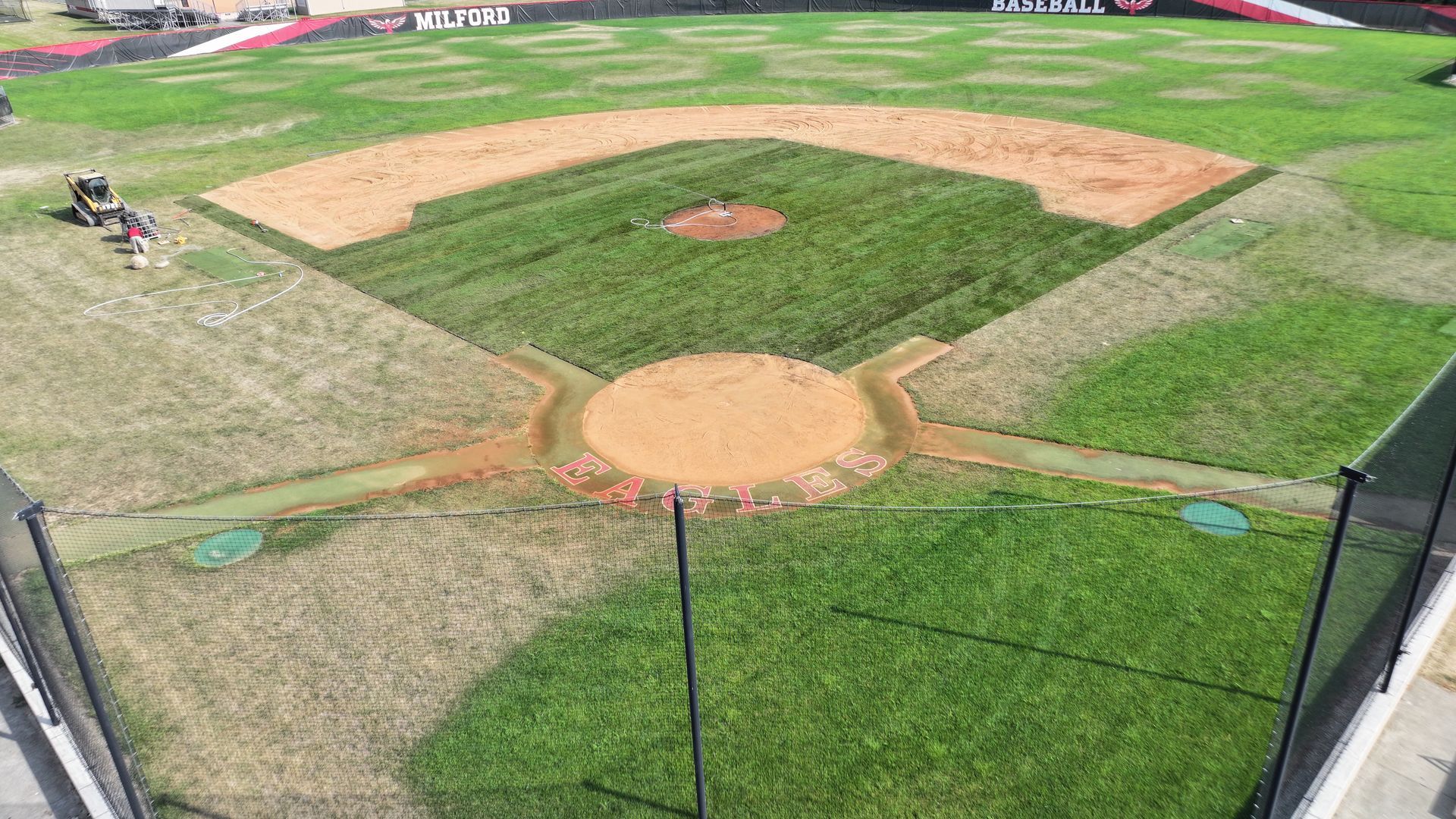 Baseball field seen from behind home plate, with dirt infield, green outfield, and a player in left field.