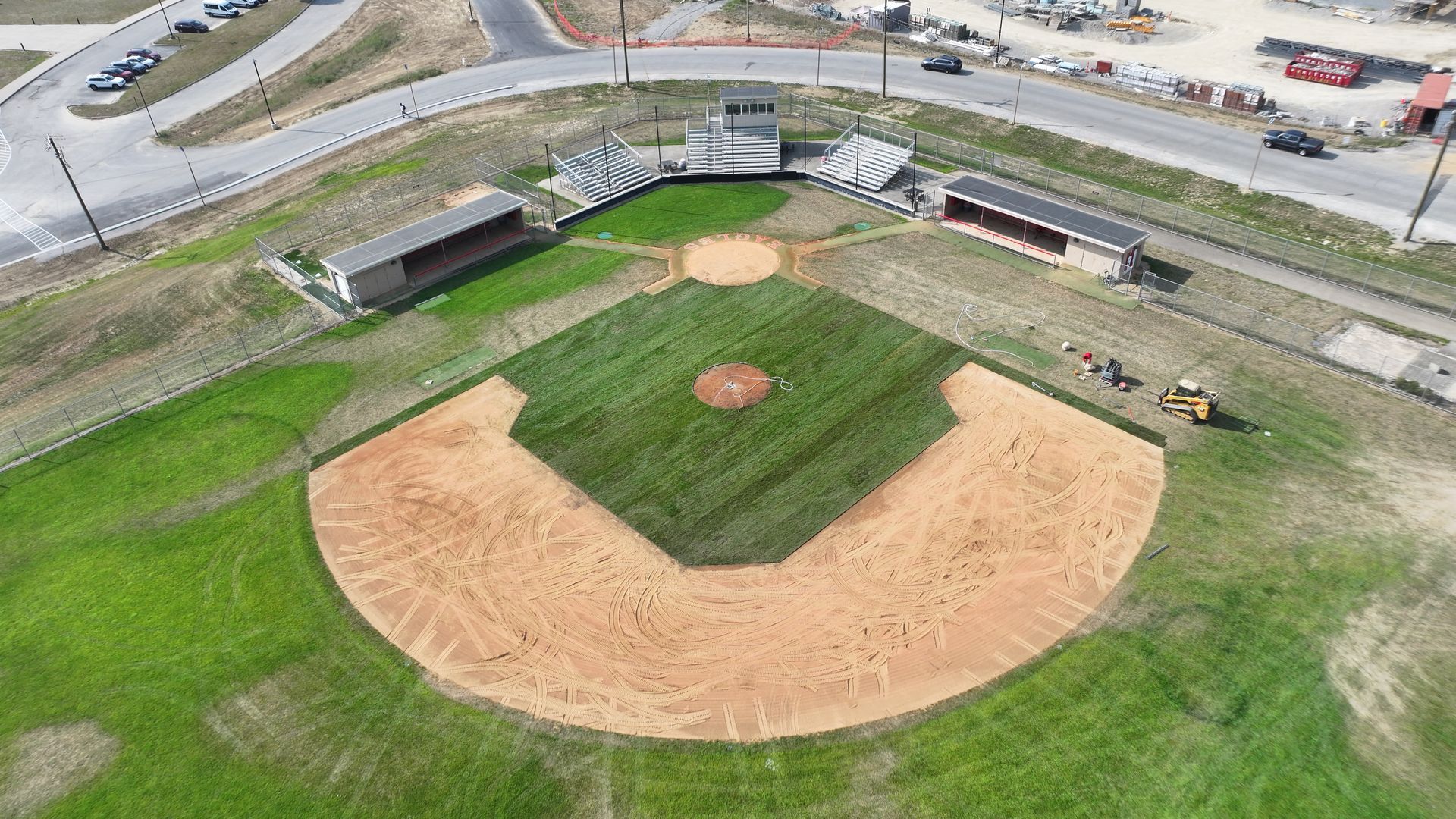 Aerial view of a small baseball field with a dirt infield, green grass, and bleachers along the outfield edges