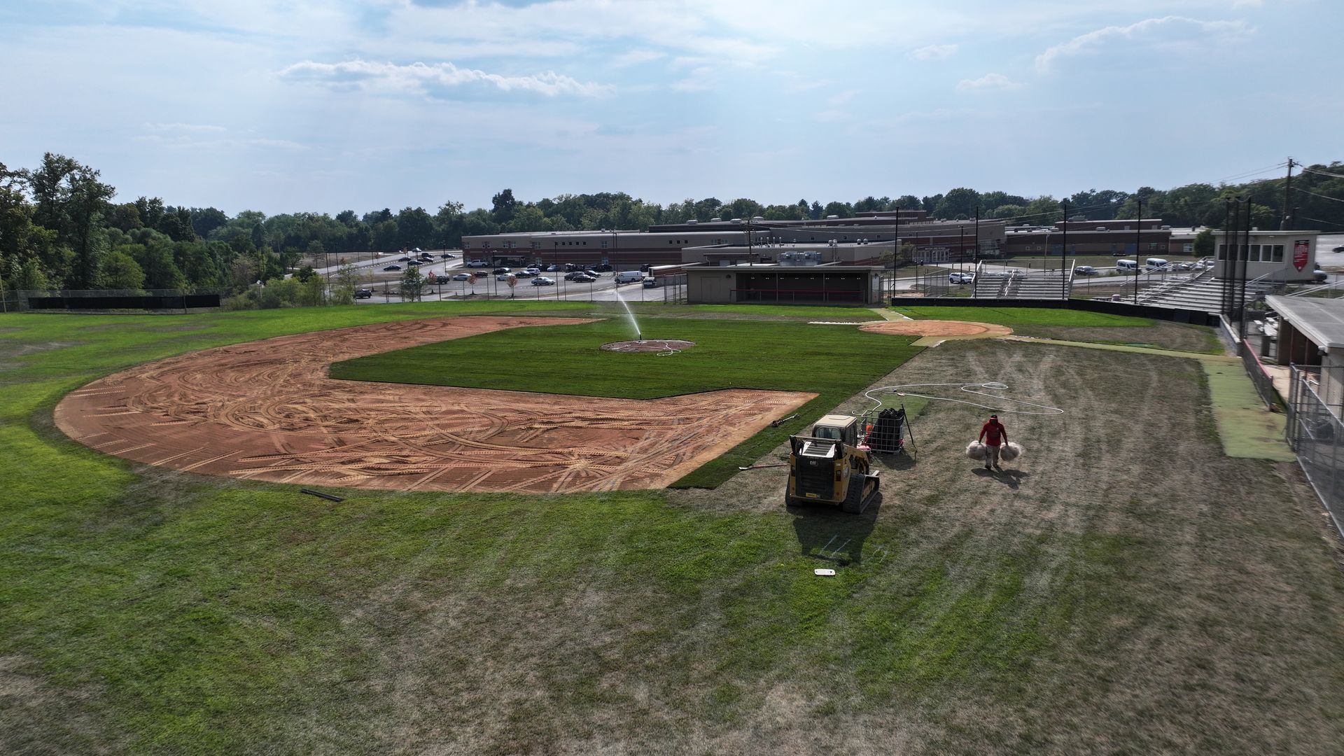 Baseball diamond under construction with excavator, workers, and green outfield under cloudy sky
