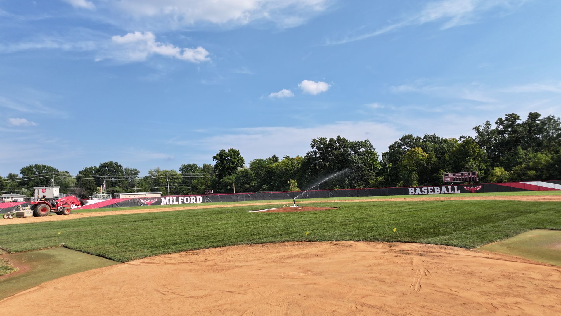 Empty dirt baseball field with green outfield under a blue sky, lined by trees and red sponsor banners