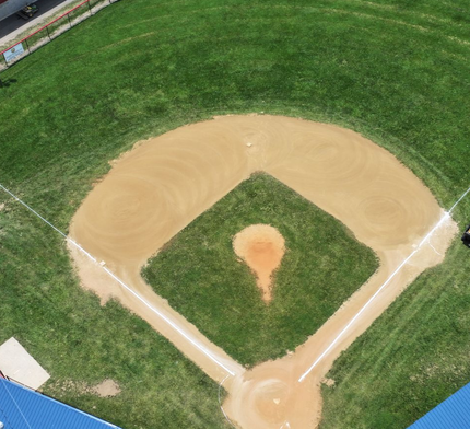Aerial view of a baseball field with a dirt infield and green outfield, centered on home plate.