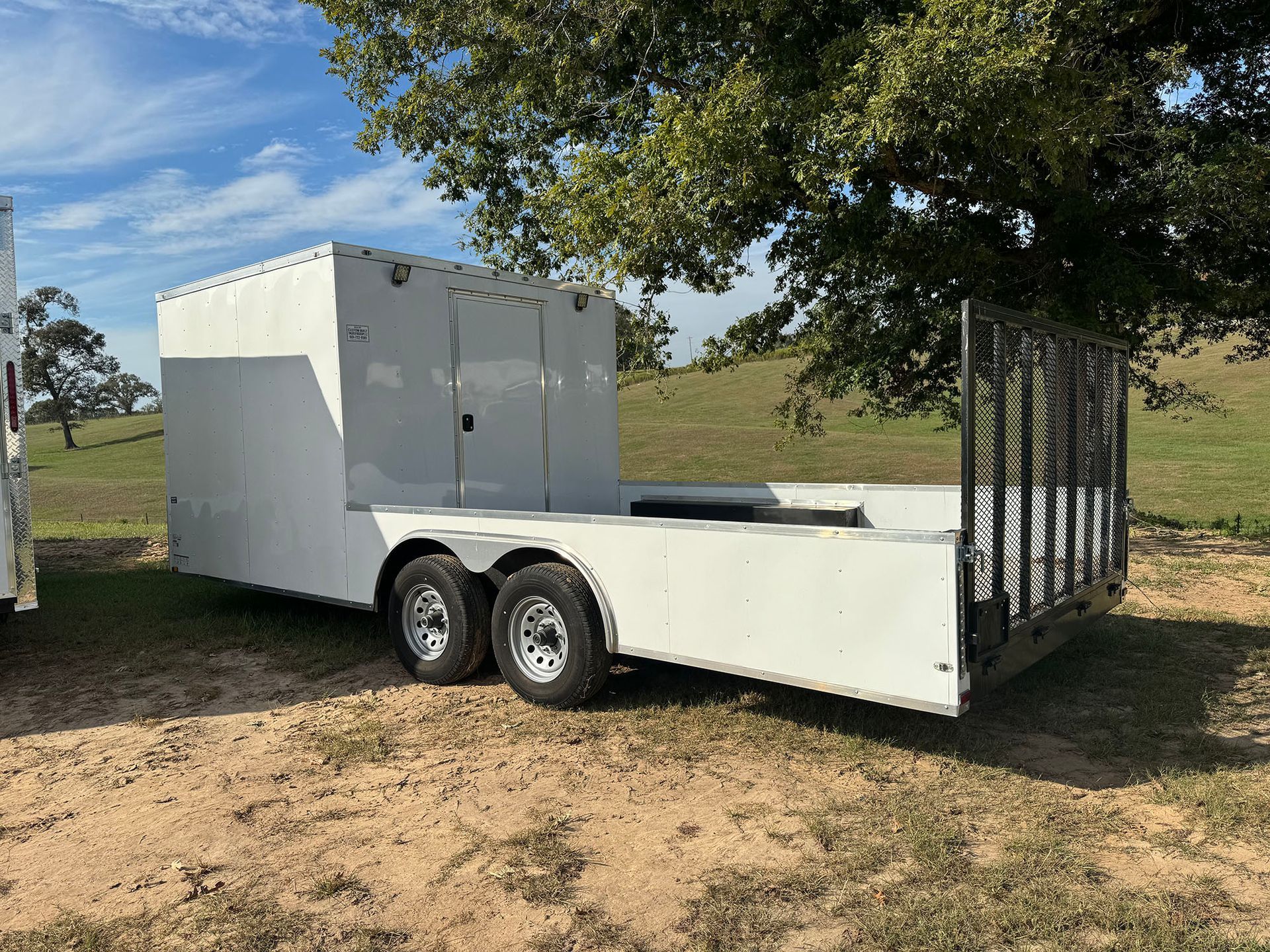 A white trailer is parked in a field under a tree.