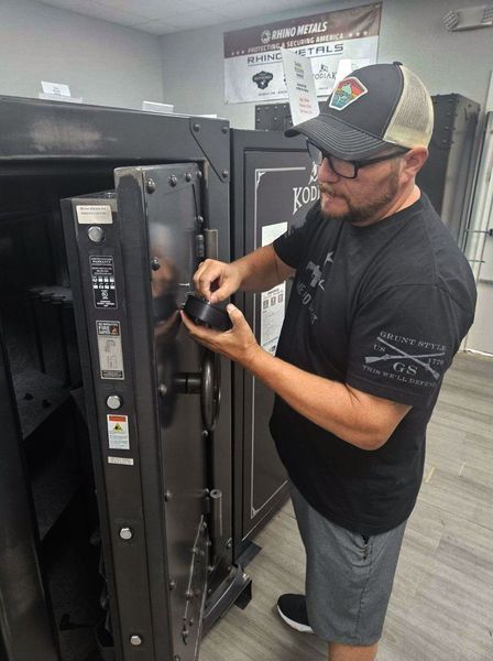 Man opening a large safe, likely in a store. He wears a hat and sunglasses, dark shirt, gray shorts, and black shoes.