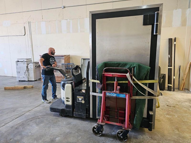 Man operates a forklift carrying a large, metal door secured with straps and a hand truck in a warehouse.