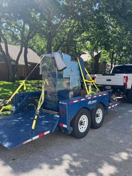 Blue trailer with a tall, silver object strapped down, parked on a street with a white truck.