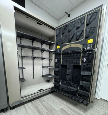 Open beige gun safe with black interior and shelves, set in a room with light wood floors.