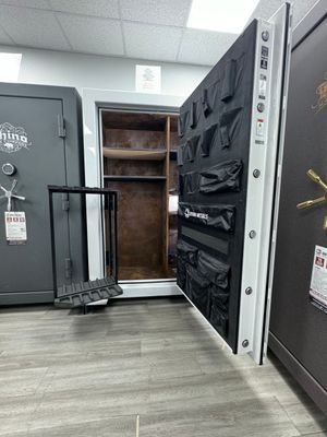 White gun safe door open, revealing interior with storage. Other safes and rack visible on showroom floor.