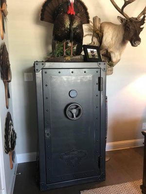 A dark grey gun safe, topped with taxidermied turkey and caribou heads, against a beige wall.