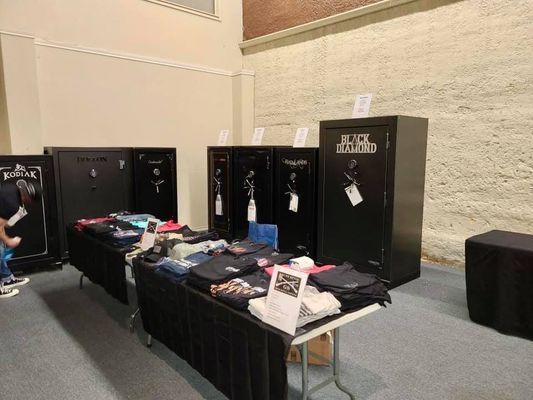 Gun safes and apparel displayed at an indoor event, featuring black safes and tables with shirts.