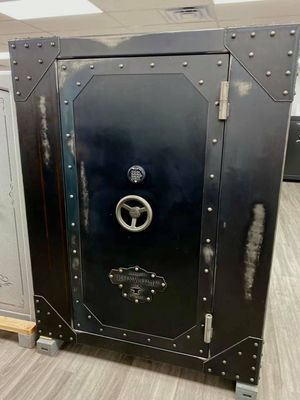 Black antique-style safe with riveted edges and combination dial, set on a gray base in a room.