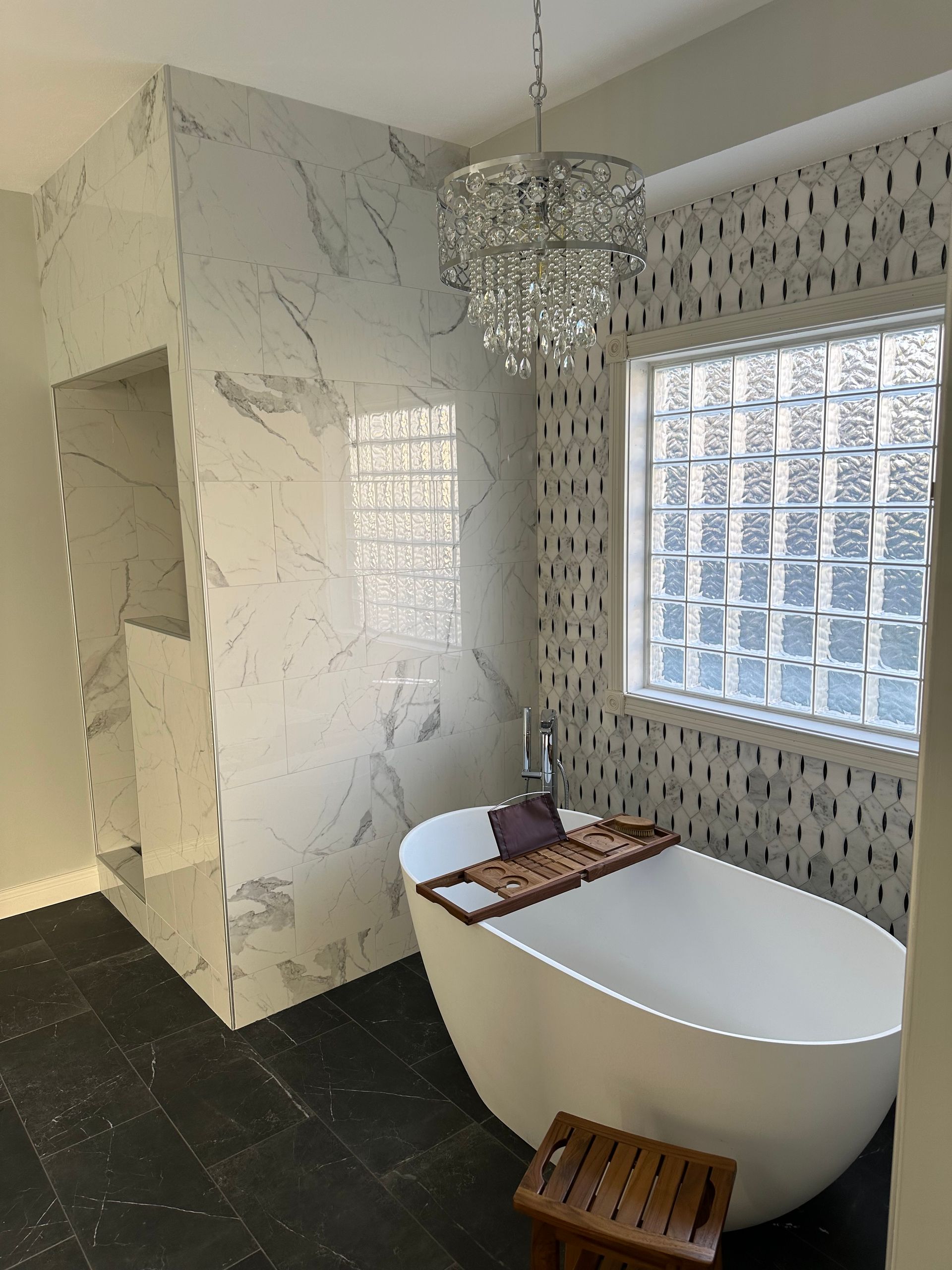 Modern bathroom with a white freestanding tub, marble walls, chandelier, and glass-block window.